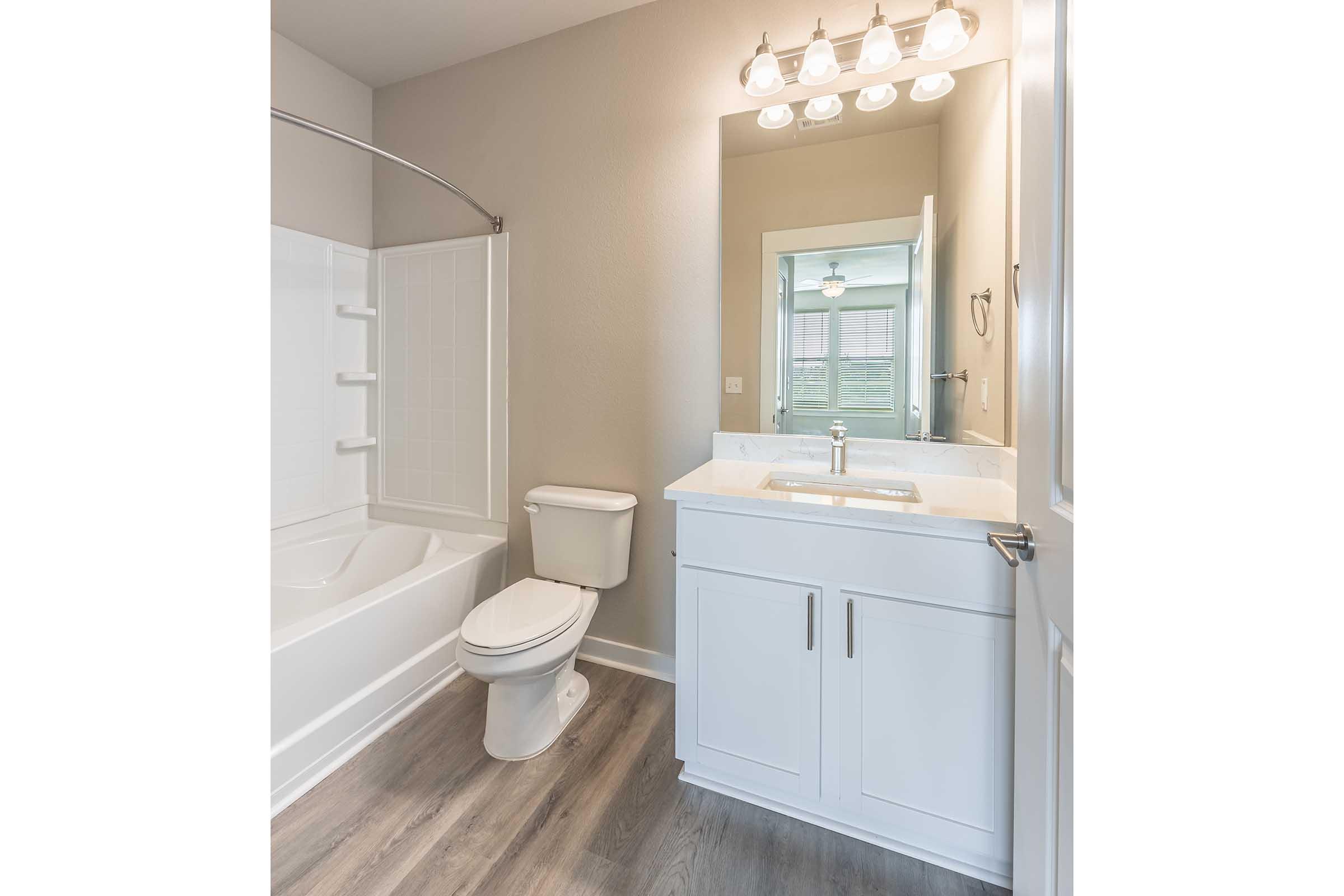 Modern bathroom featuring a white bathtub, toilet, and a vanity with a sink. The space has neutral-colored walls, a large mirror above the sink, and a bright overhead light fixture. A window is visible in the background, allowing natural light to enhance the room's airy feel.