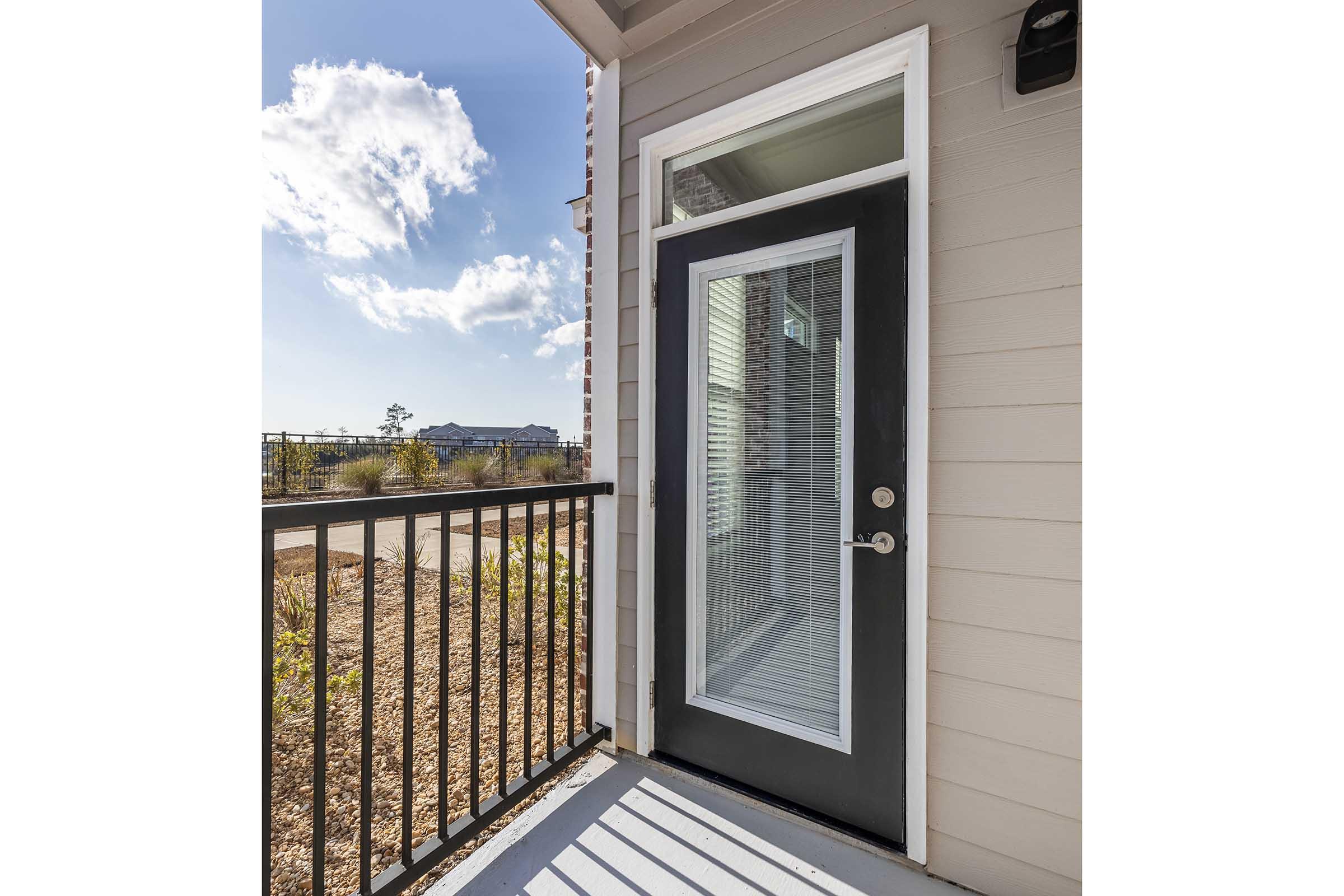 Exterior view of a modern front door with a large glass panel, framed in a sleek black finish. The door is set against a light-colored wall. Sunlight casts shadows on the porch, which features a railing. In the background, there are glimpses of a neighborhood and a blue sky with clouds.