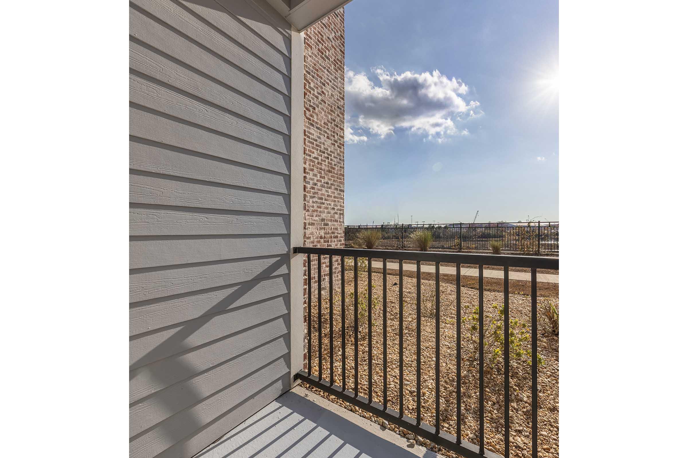 A corner balcony with a metal railing, featuring a view of a wide landscape under a blue sky with fluffy clouds and sunlight. The balcony is surrounded by light gray siding and exposed brick, providing a modern aesthetic. The ground below is covered with gravel and sparse vegetation.