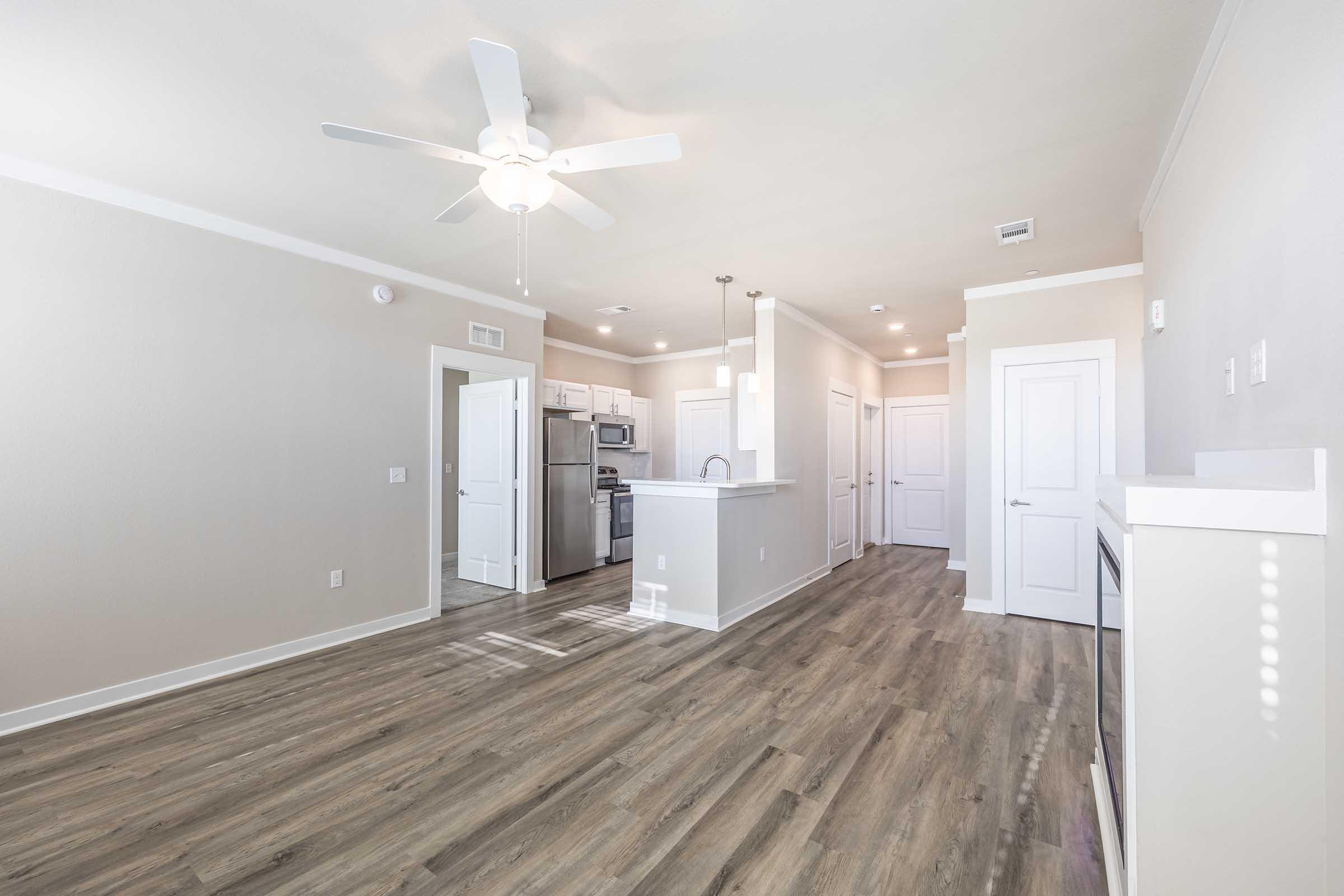 Interior view of a modern apartment showcasing an open floor plan. The space features light-colored walls, a ceiling fan, and laminate flooring. A kitchen area is visible with stainless steel appliances and a small island. Multiple doors lead to other rooms, creating a bright and airy atmosphere.