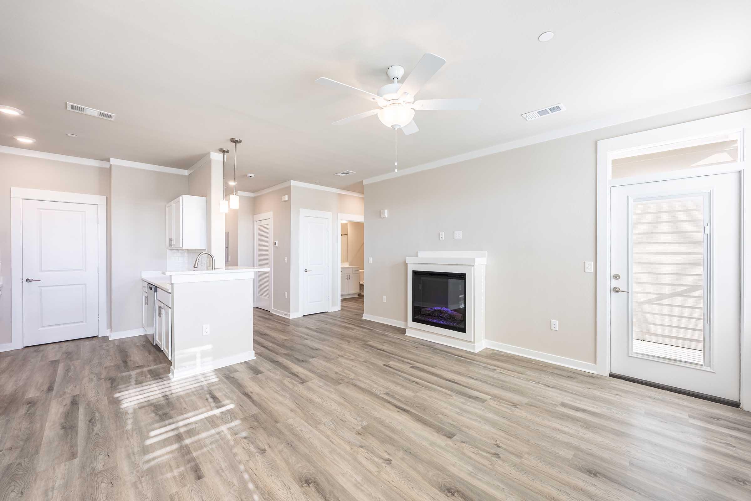 Bright and spacious living area featuring modern decor, a ceiling fan, wood-like flooring, and a cozy electric fireplace. The open layout includes a kitchen area with white cabinetry and access to a doorway leading outside. Natural light flows in through large windows, creating a welcoming atmosphere.