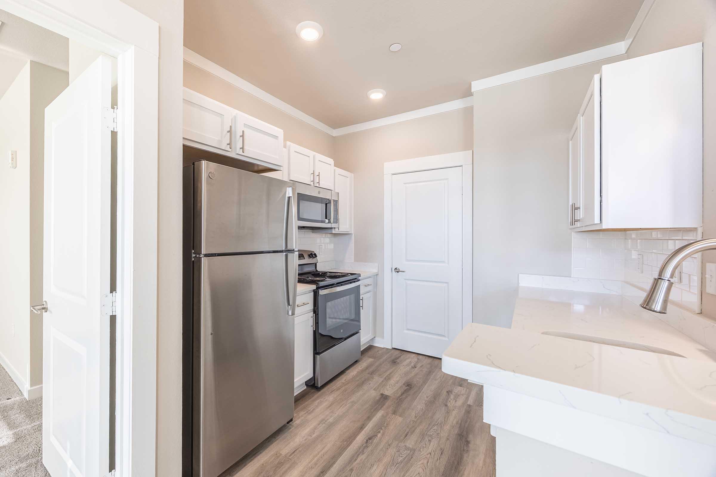 A modern kitchen featuring stainless steel appliances, including a fridge, oven, and microwave. White cabinetry and a countertop with a sink are visible. The walls are painted a light beige color, and there is natural light coming from an adjacent room. The floor is covered with wood-like laminate.