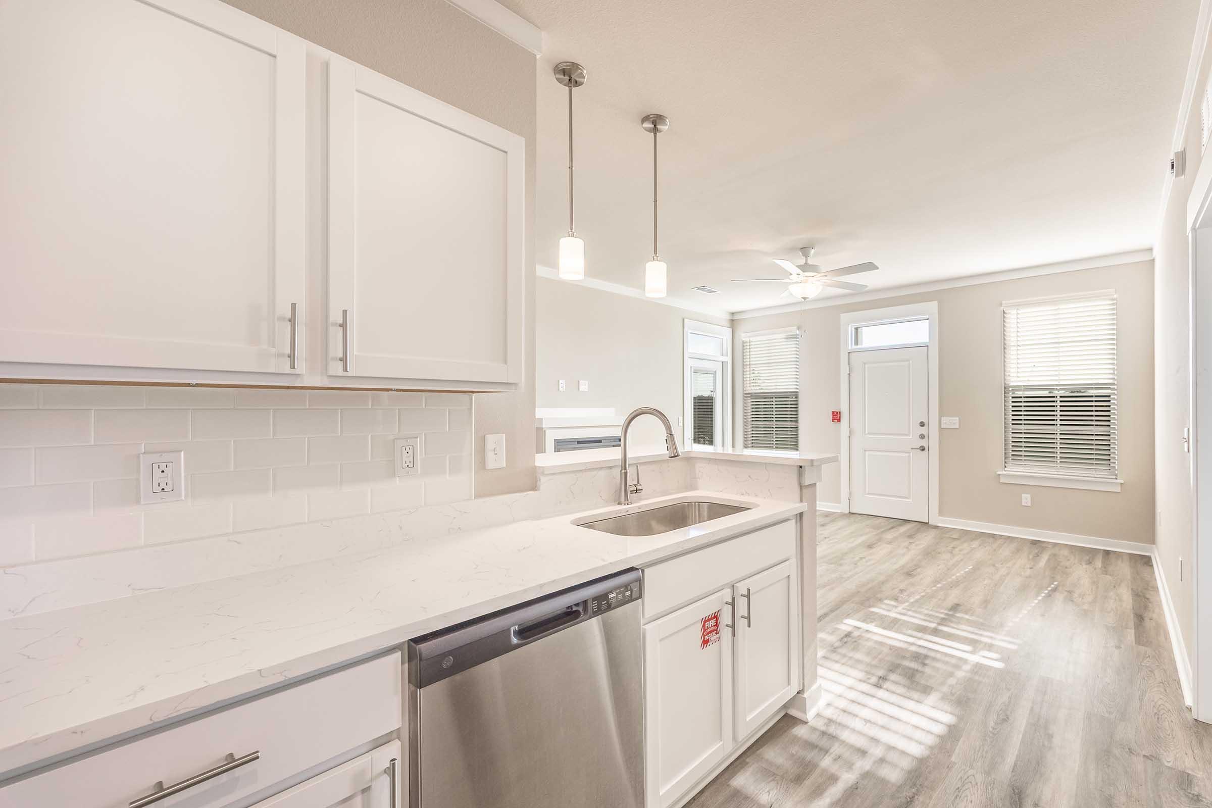Modern kitchen interior featuring white cabinetry, stainless steel appliances, and a quartz countertop. The space is well-lit with natural light coming from nearby windows. There is a dining area visible in the background, and the flooring is a light-colored wood laminate.