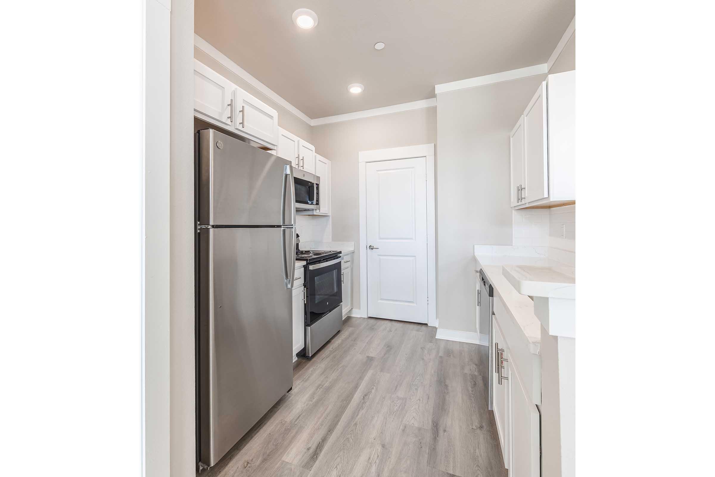A modern kitchen featuring stainless steel appliances, including a refrigerator, oven, and microwave. The cabinetry is white with a light backsplash, and the floor is a light wood laminate. The kitchen has recessed lighting and an open layout with a door leading to another room.