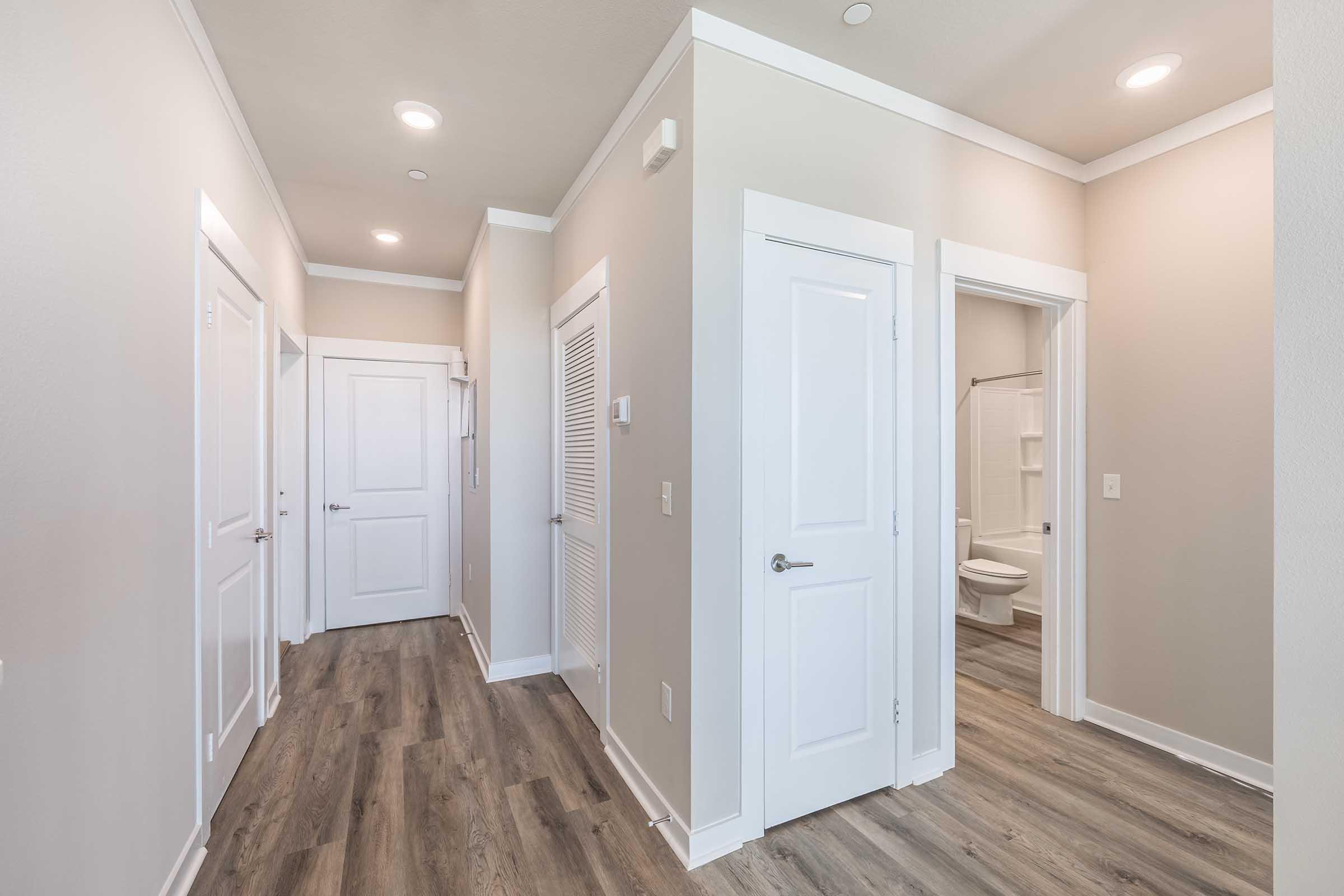 A well-lit hallway featuring light-colored walls and modern laminate flooring. There are two closed doors on the left, one with a vent, and an open door on the right leading to a bathroom. The ceiling is equipped with recessed lighting, creating a bright and spacious atmosphere.