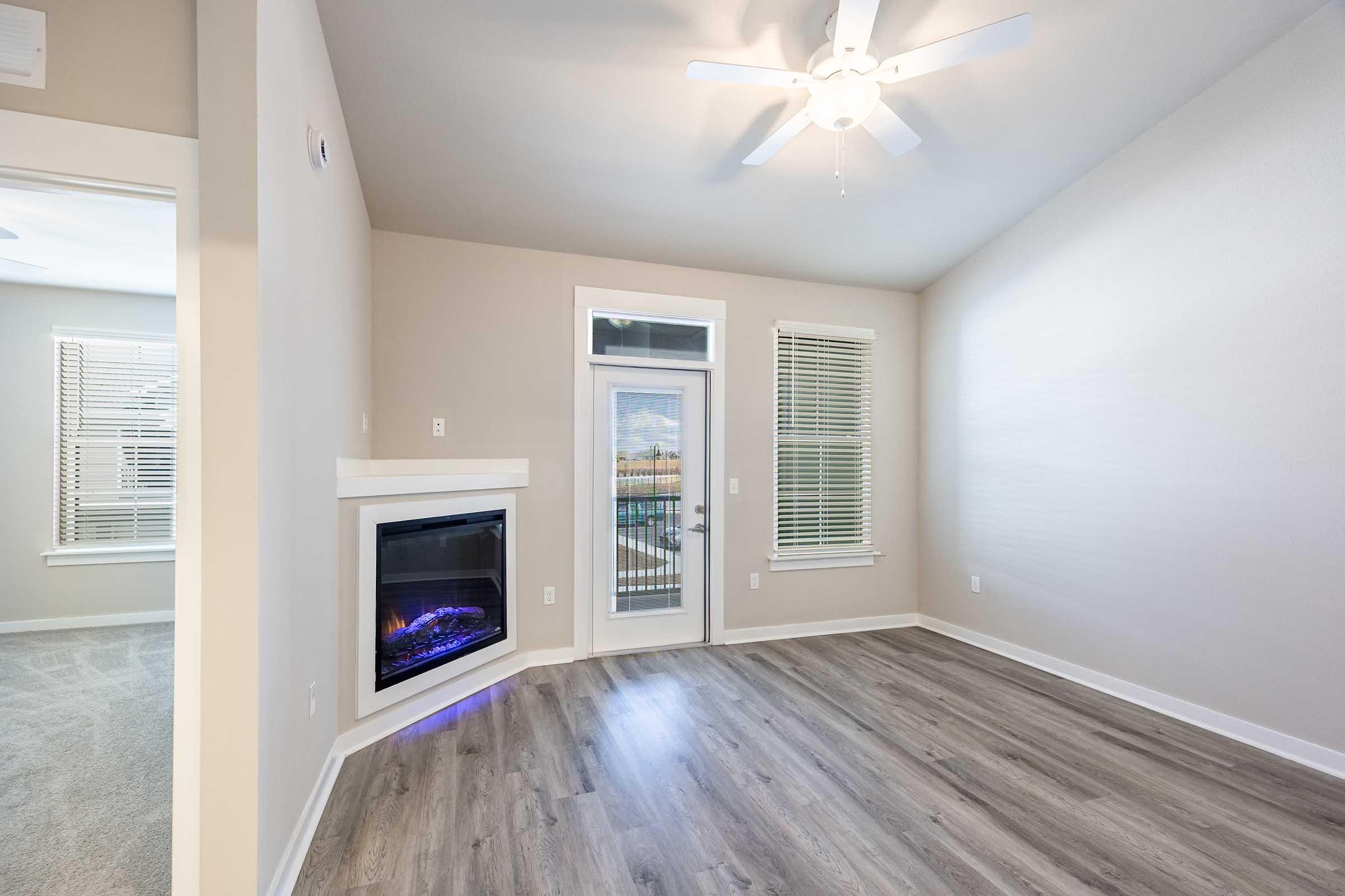 A bright, spacious room with light-colored walls and hardwood flooring. It features a modern electric fireplace below a flat-screen TV mount, a ceiling fan, and large windows with blinds. The room opens to a glass door leading outside, providing natural light and a view.