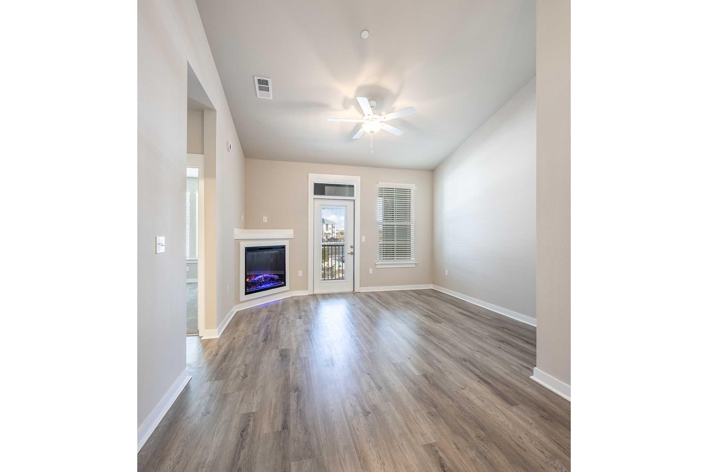 Spacious living room with light-colored walls and hardwood flooring, featuring a ceiling fan and a cozy electric fireplace. Large windows allow natural light, and there's a balcony visible through the glass door. The overall atmosphere is bright and inviting.