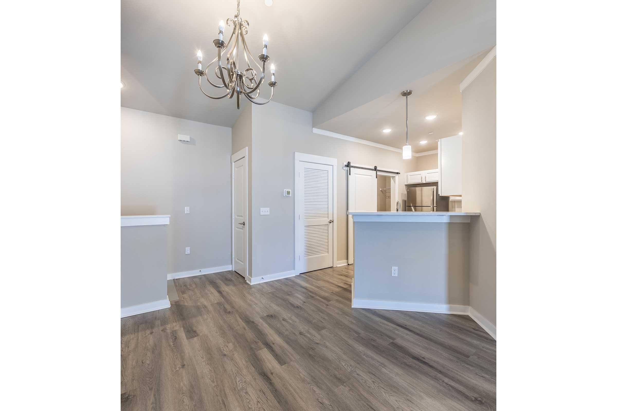 An open-concept living space featuring a modern chandelier, light gray walls, and new laminate flooring. A kitchen area with bar seating is visible in the background, along with a closed door leading to another room. The overall ambiance is bright and contemporary, ideal for a cozy living environment.