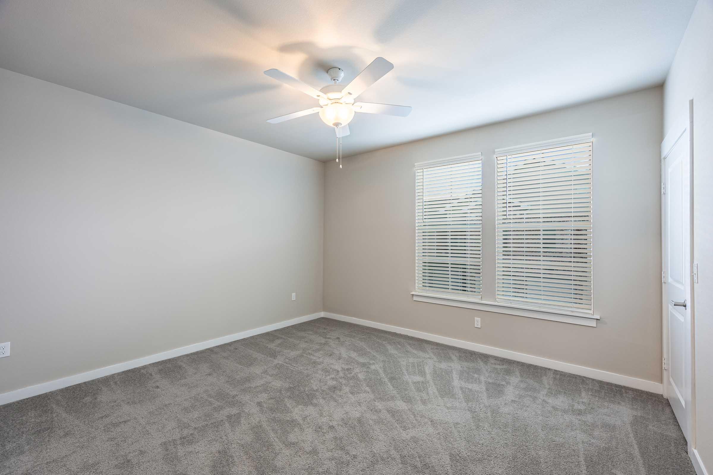 Empty room featuring light gray walls and carpet, a ceiling fan, and two windows with white blinds allowing natural light. The space is uncluttered and ready for furnishing, with a door leading to another area.