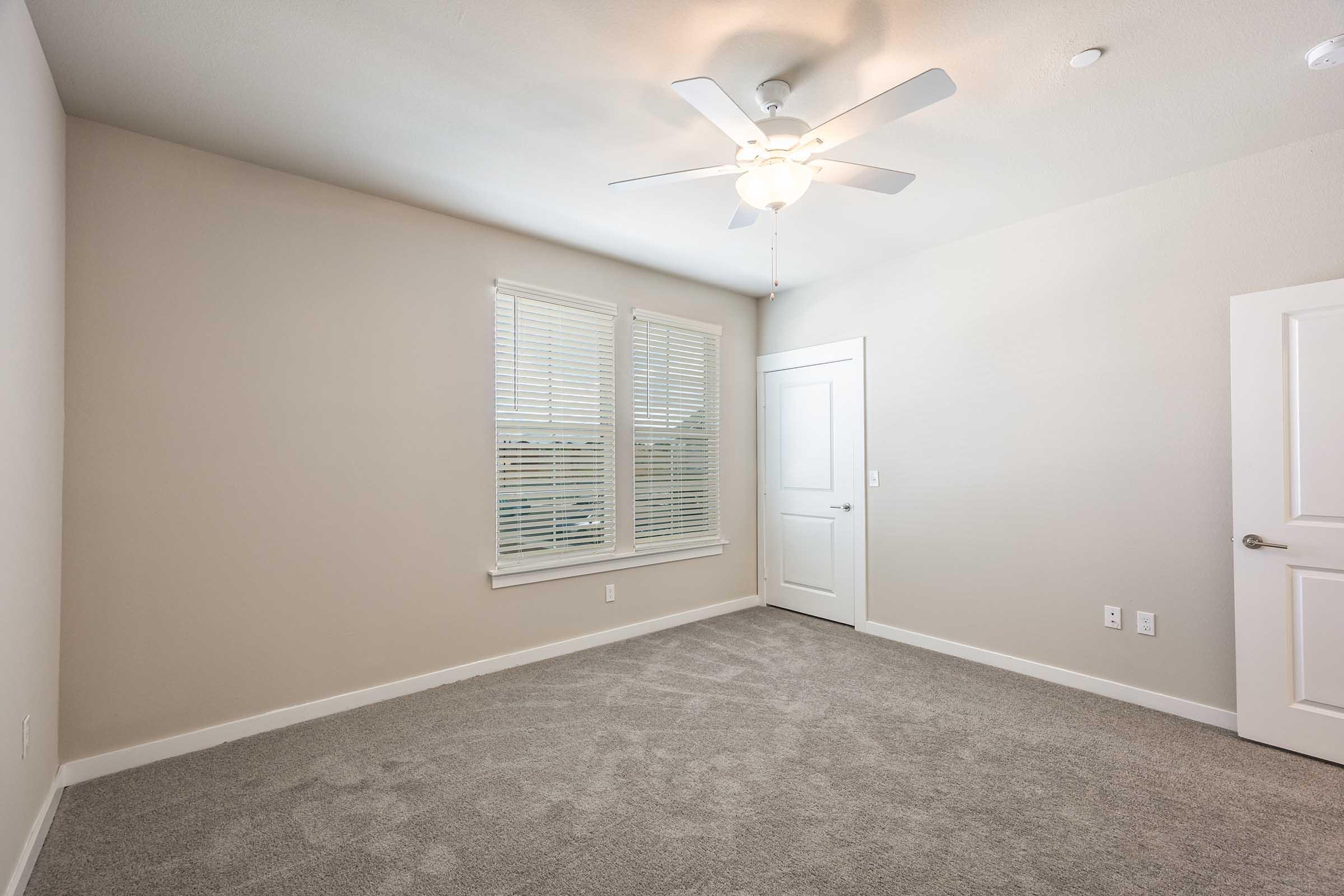 A blank room with light beige walls and grey carpeting. It features a ceiling fan, two windows with white blinds, and a closed white door. The space is empty, providing a clean and spacious look, suitable for various configurations or furnishings.