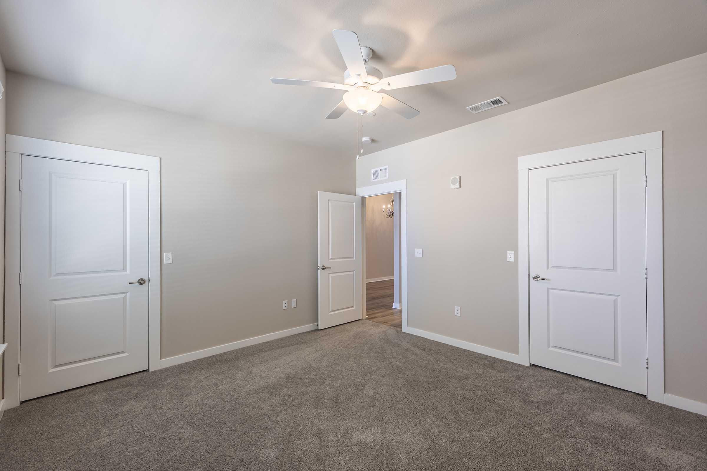 A bright and spacious room featuring light beige walls and plush carpet flooring. It has a ceiling fan and two white doors on opposite walls, leading to other areas. Natural light enters, creating a warm and inviting atmosphere. The room is empty and ready for decoration.