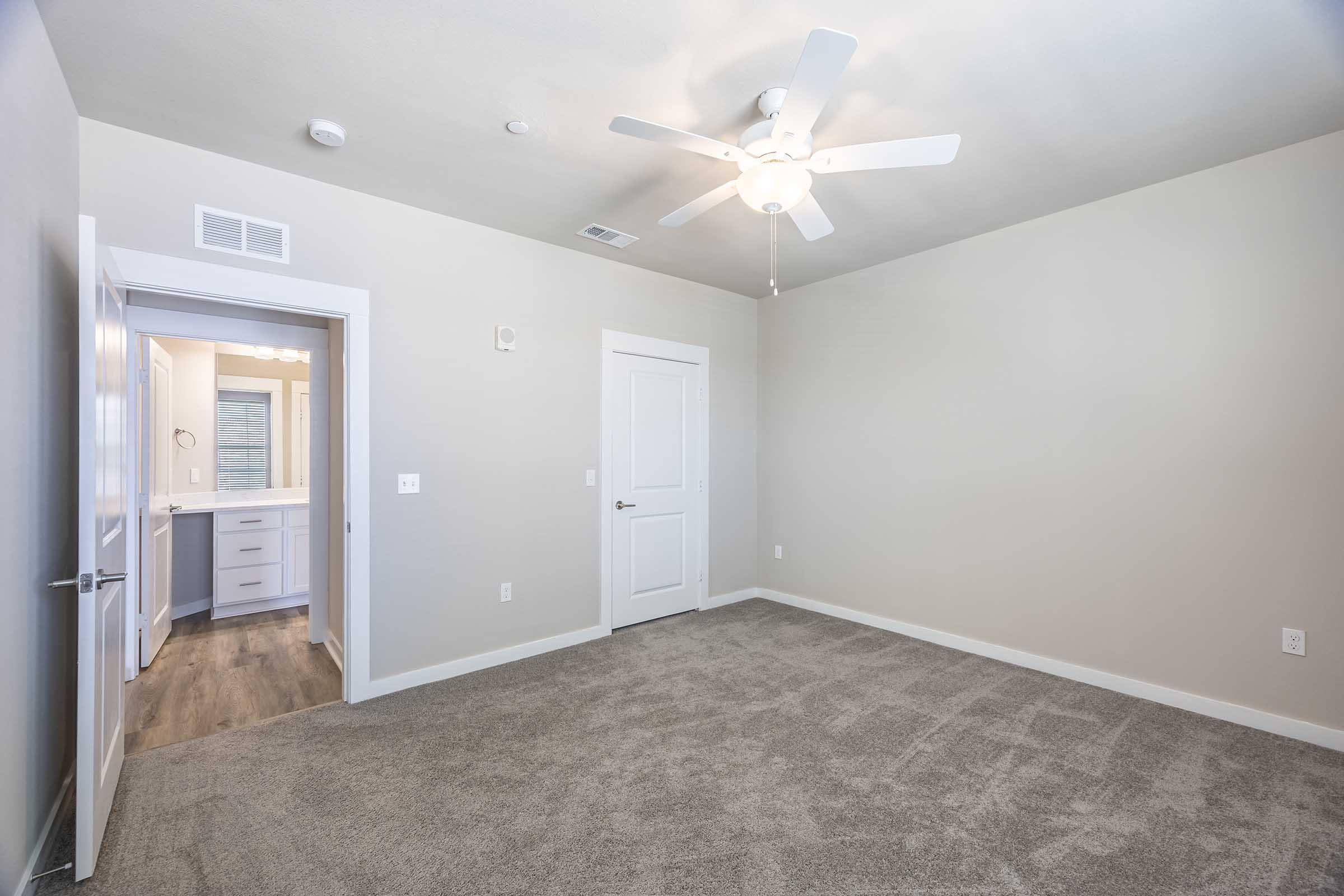 A spacious, empty bedroom featuring light gray walls and carpet. A ceiling fan is installed, and there’s a doorway leading to a bathroom. Natural light enters from the door, enhancing the open feel of the room.