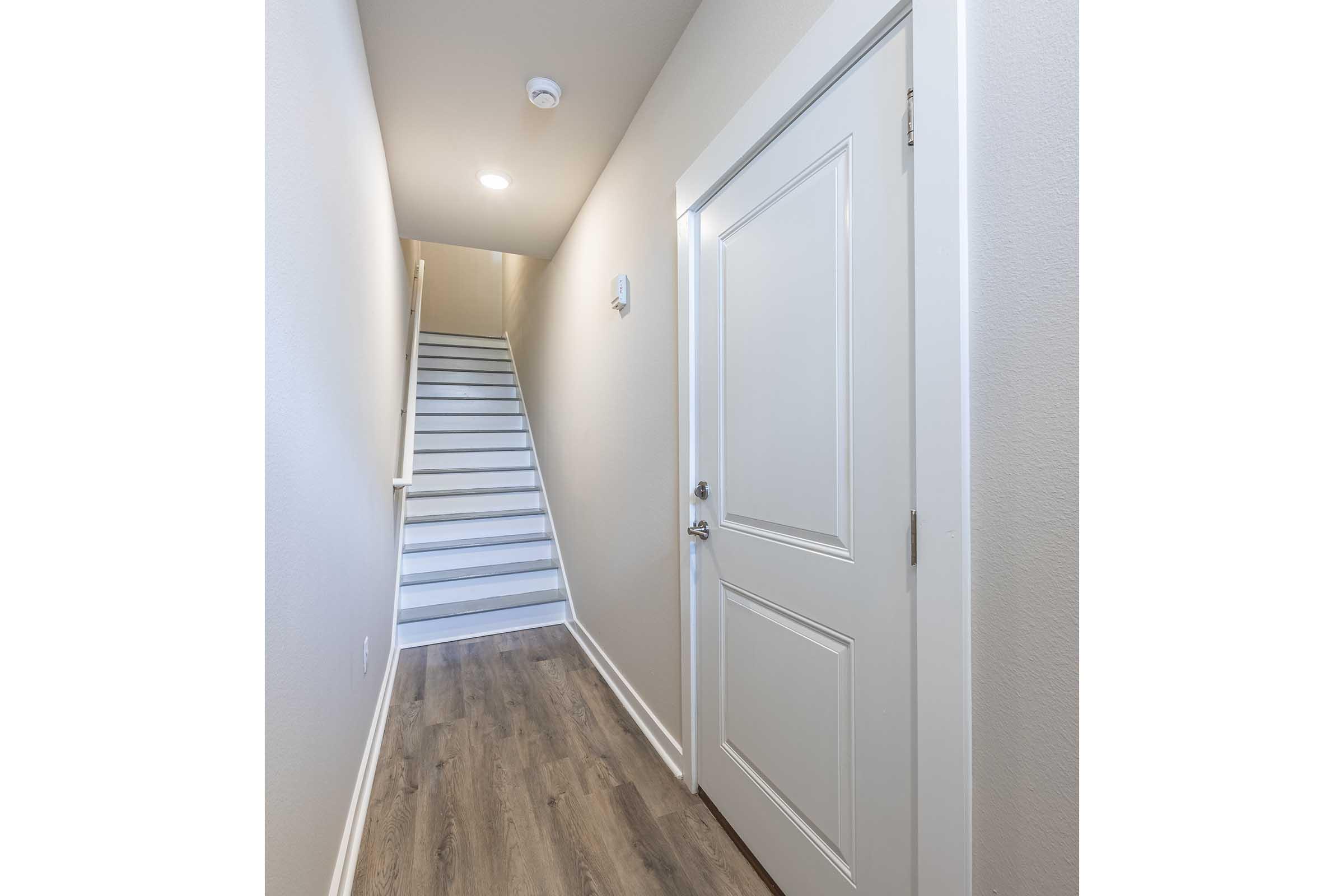 A well-lit hallway featuring a white door on the left and a staircase ahead leading to the upper level. The walls are painted in a light neutral color, and the flooring is a wooden laminate. The space is uncluttered and modern, conveying a sense of simplicity and freshness.