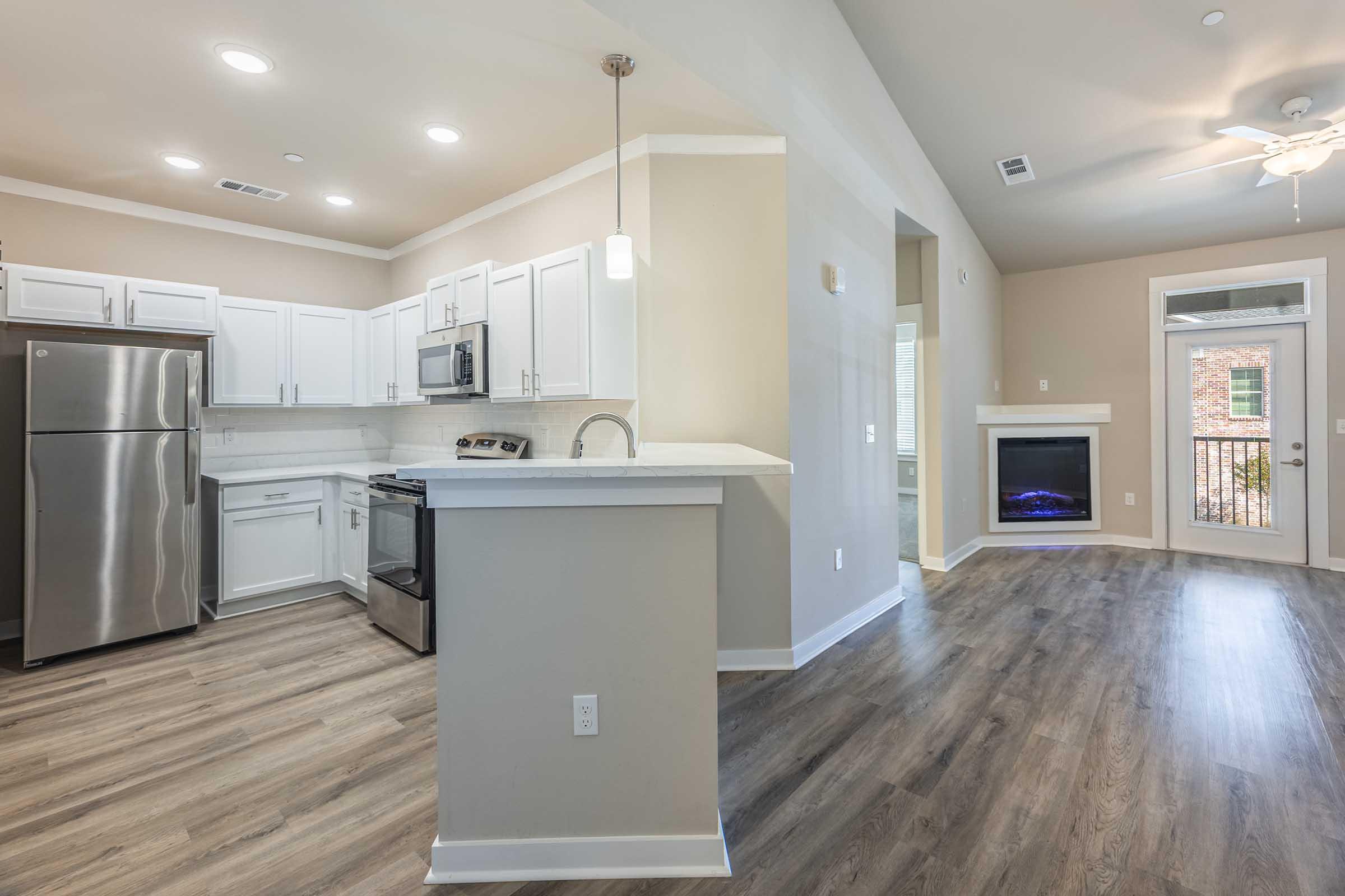 Modern kitchen and living area featuring stainless steel appliances, white cabinetry, and a gray countertop. The kitchen opens to a spacious living room with a fireplace and large windows allowing natural light. The flooring is a light wood laminate, creating a warm and inviting atmosphere.