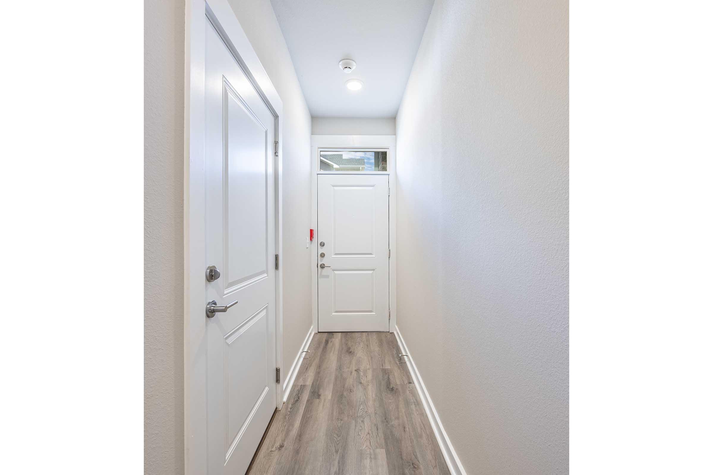 A narrow hallway featuring white walls and a light wood floor. At the end of the hallway, there is a white door with a window above it. A small ceiling light illuminates the space, creating a bright and welcoming atmosphere.