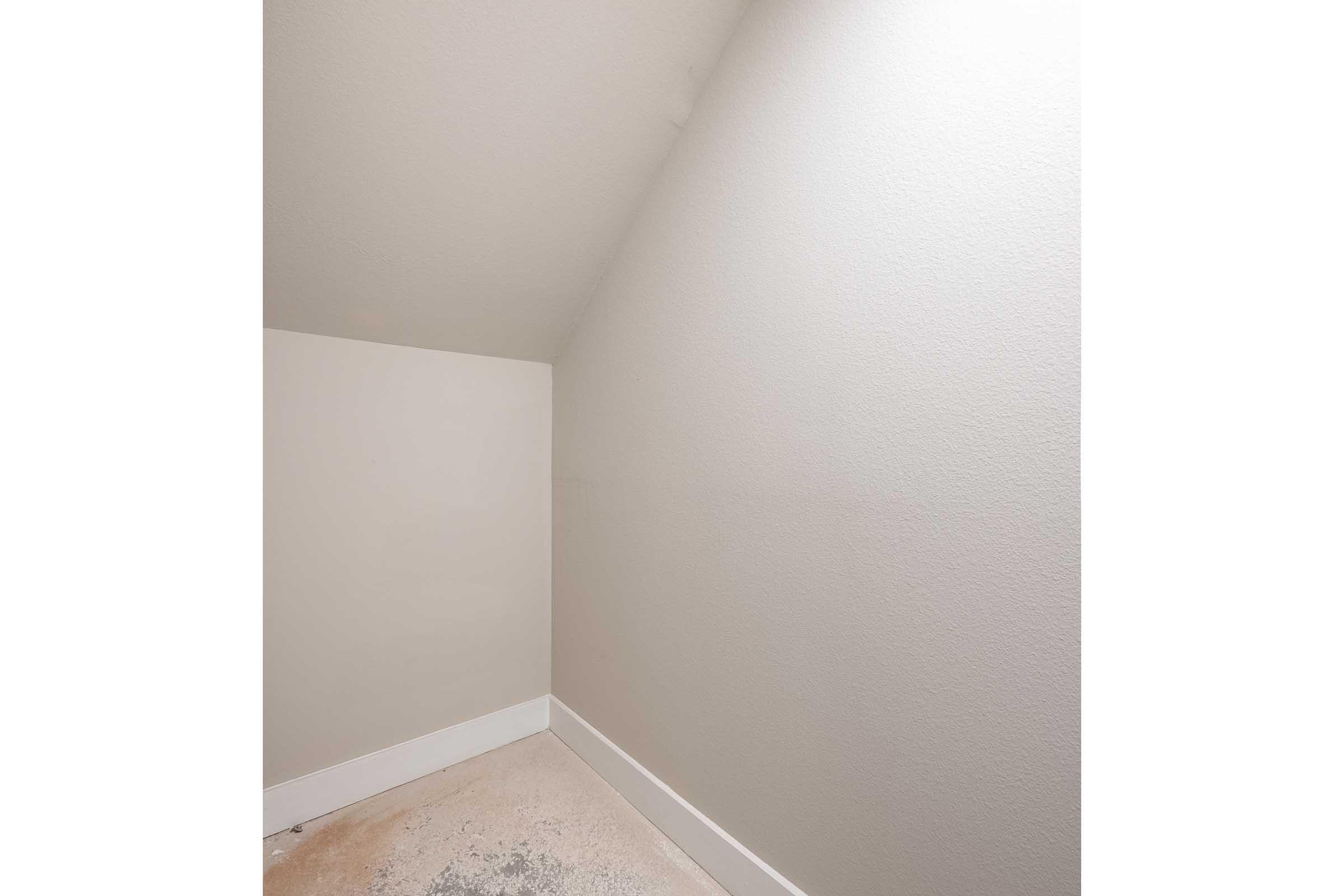 A small, unfinished attic space with sloped walls and a light gray color. The floor is partially covered with dust and debris, while the corners of the room are well-defined. The overall atmosphere is simple and minimalistic, with no furniture or decorations present.