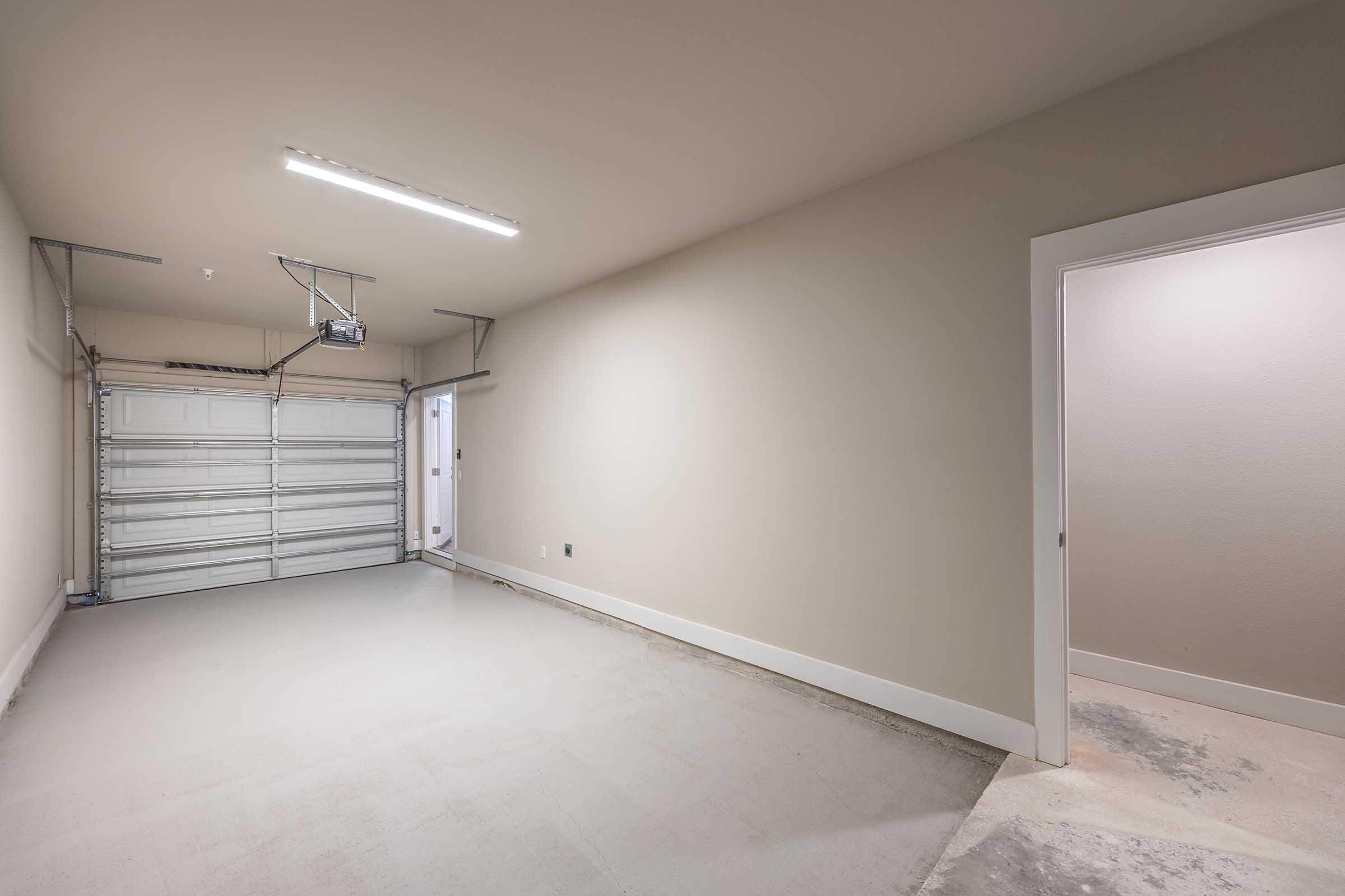 Interior of a garage featuring a garage door on the left, a side door on the right, and a clean, empty concrete floor. The walls are painted a neutral color with bright overhead lighting.