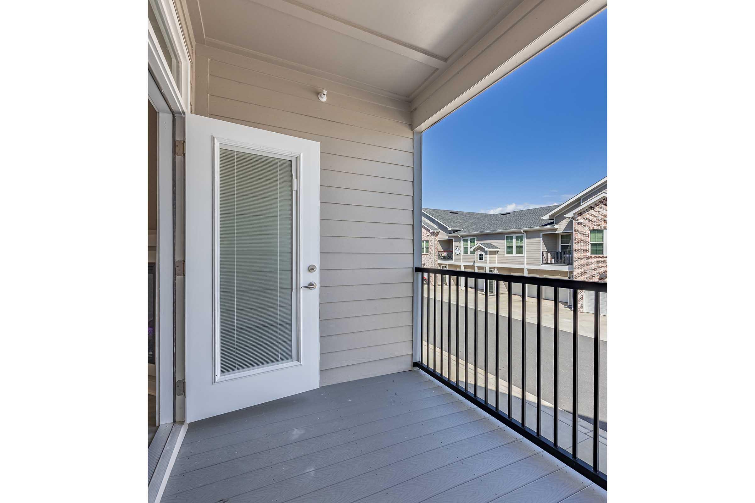 A balcony area featuring gray flooring and light-colored siding. A glass door leads into an interior space, and the view shows residential buildings and blue sky in the background. The railing is black, adding a modern touch to the outdoor area.
