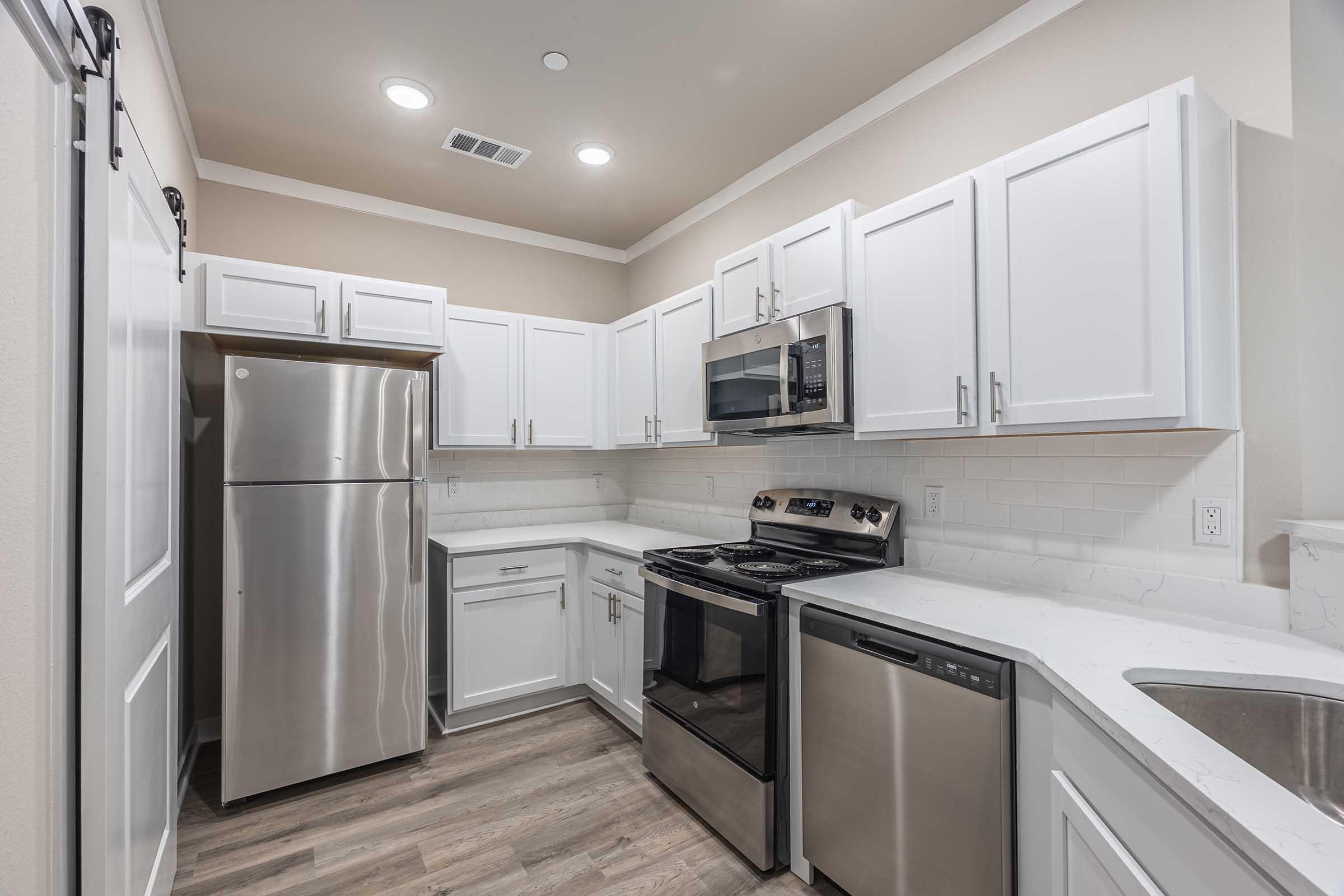 Modern kitchen featuring stainless steel appliances, including a refrigerator, stove, and dishwasher. The cabinetry is white with black handles, and there is a spacious countertop. Neutral-colored walls and wood-look flooring complete the contemporary look. Soft lighting illuminates the space.