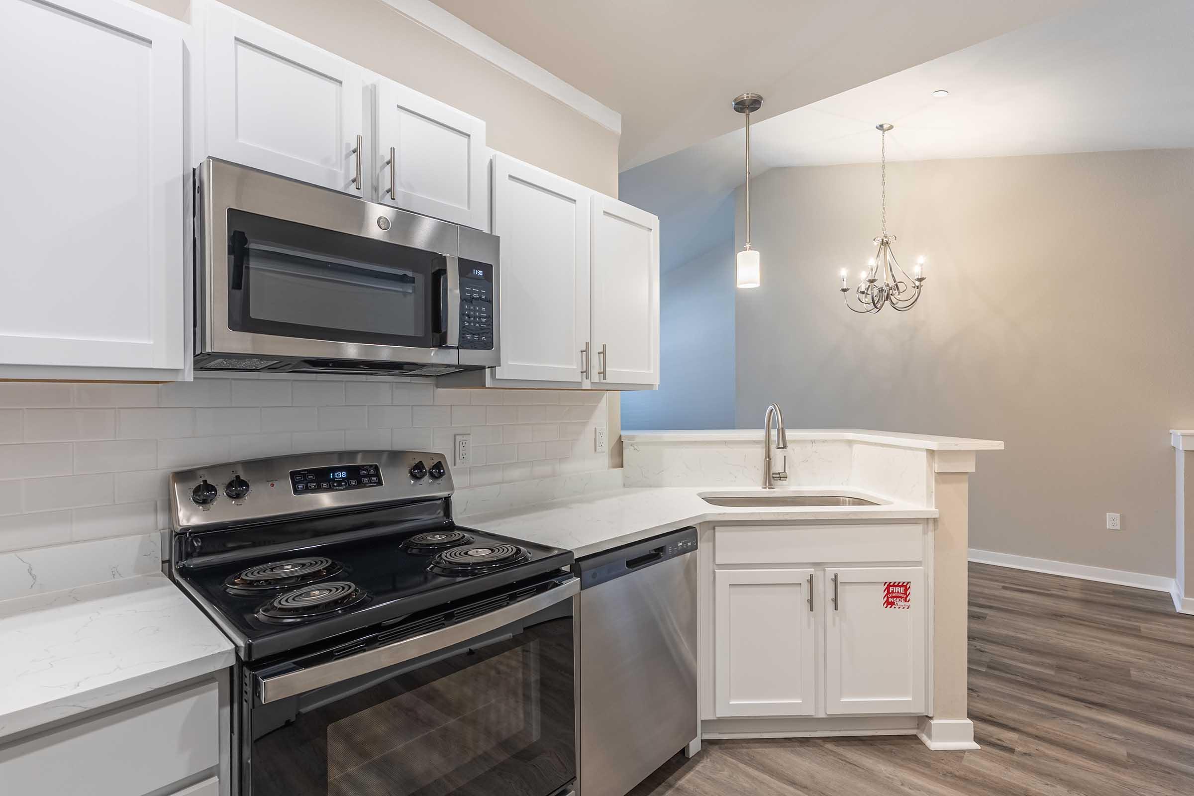 Modern kitchen featuring stainless steel appliances, including a microwave and stove, white cabinetry, and a sleek countertop. The design includes a double bowl sink with a modern faucet and pendant lighting above. The flooring is a light wood finish, creating a bright and inviting atmosphere.