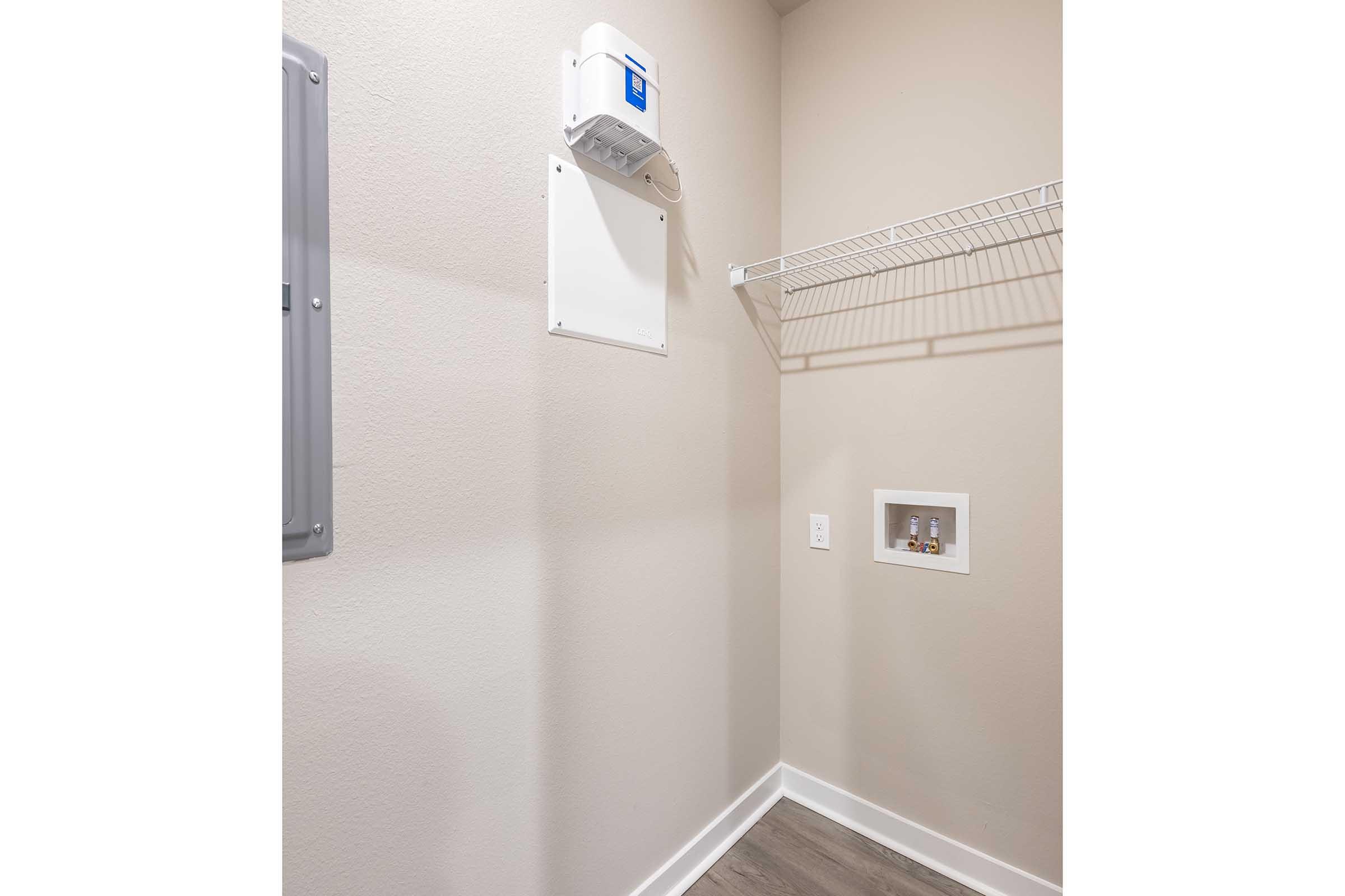 A small, empty laundry room featuring a light-colored wall, a wire shelf mounted above, and a panel on the left side. There is a power outlet and space for appliances. The floor has a light wood-like appearance.