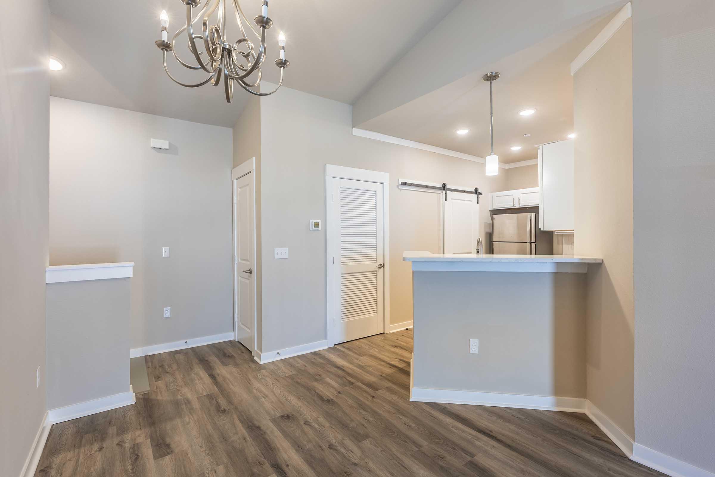 A modern kitchen and living area featuring a chandelier, light-colored walls, and wood-style flooring. The kitchen has a breakfast bar, stainless steel appliances, and a door leading to another room. Natural light fills the space, creating a warm and inviting atmosphere.