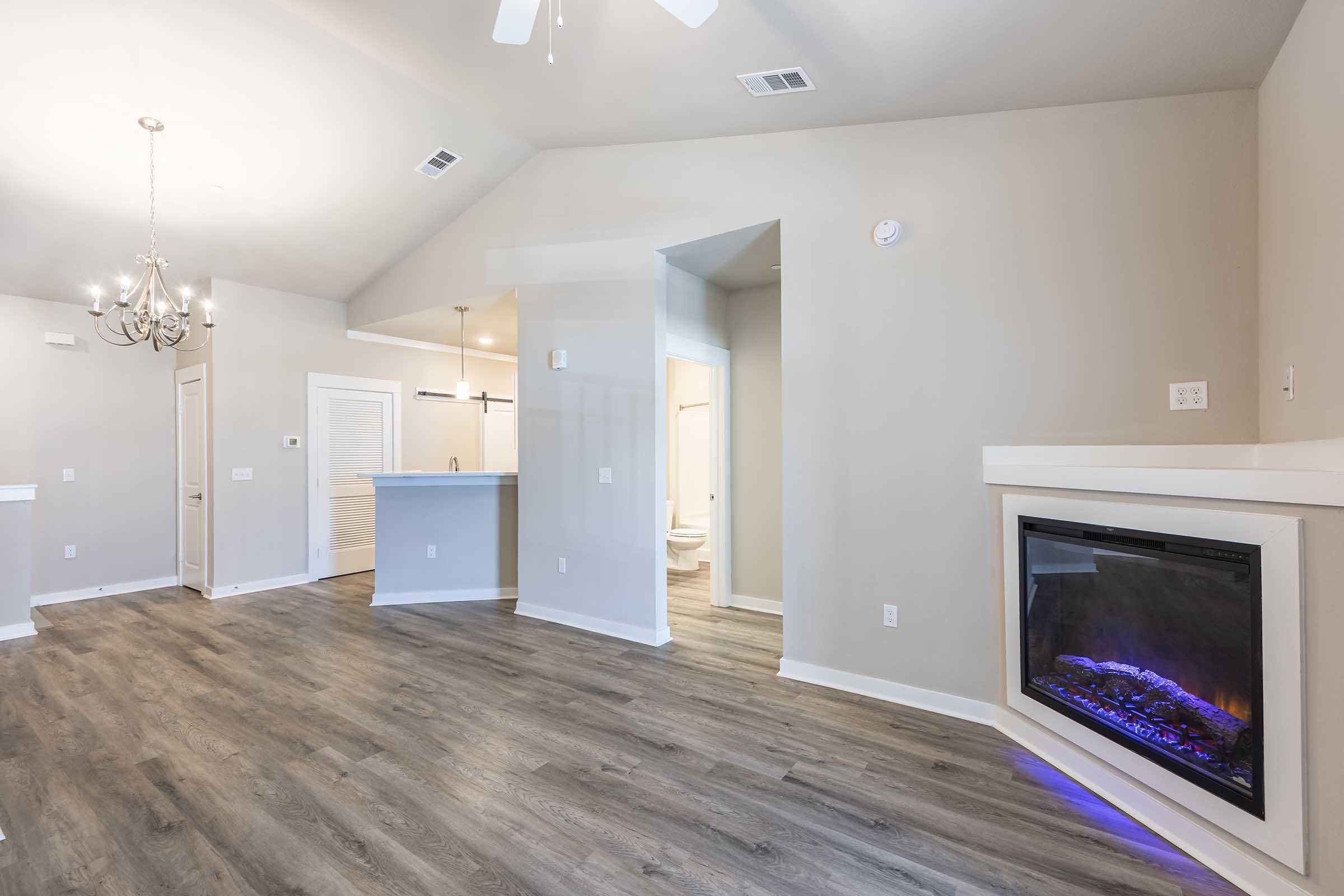 A modern living area featuring an open layout with light gray walls and wood-like laminate flooring. On the right, there is a sleek electric fireplace, and to the left, a dining area with a chandelier. The kitchen is visible in the background with white cabinetry. Natural light brightens the room, creating a warm atmosphere.