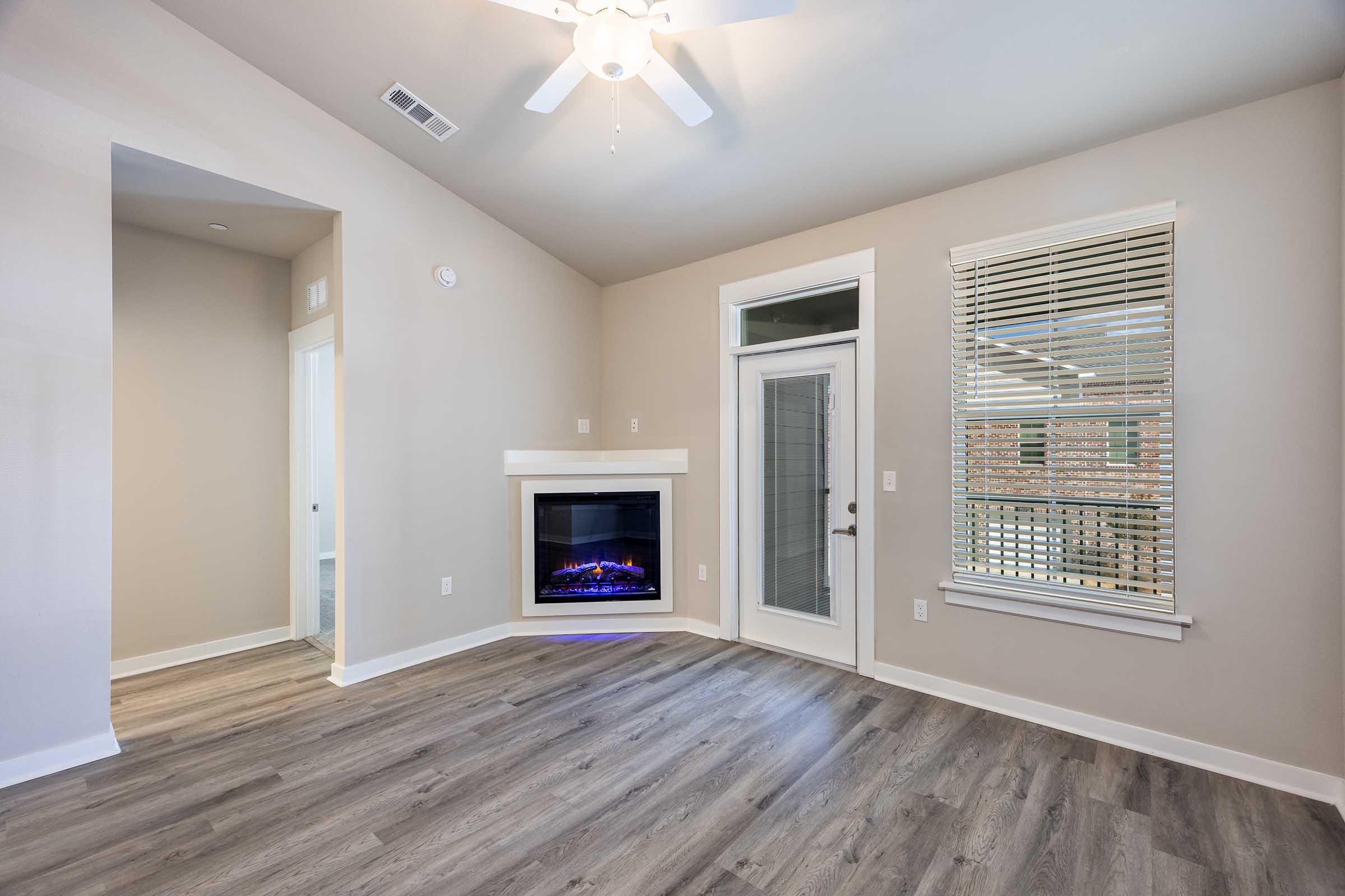 Bright living room featuring a modern electric fireplace, light gray walls, and large windows with blinds. The floor is covered in light-colored laminate, creating a spacious and inviting atmosphere. A ceiling fan adds comfort, and a doorway leads to another room.