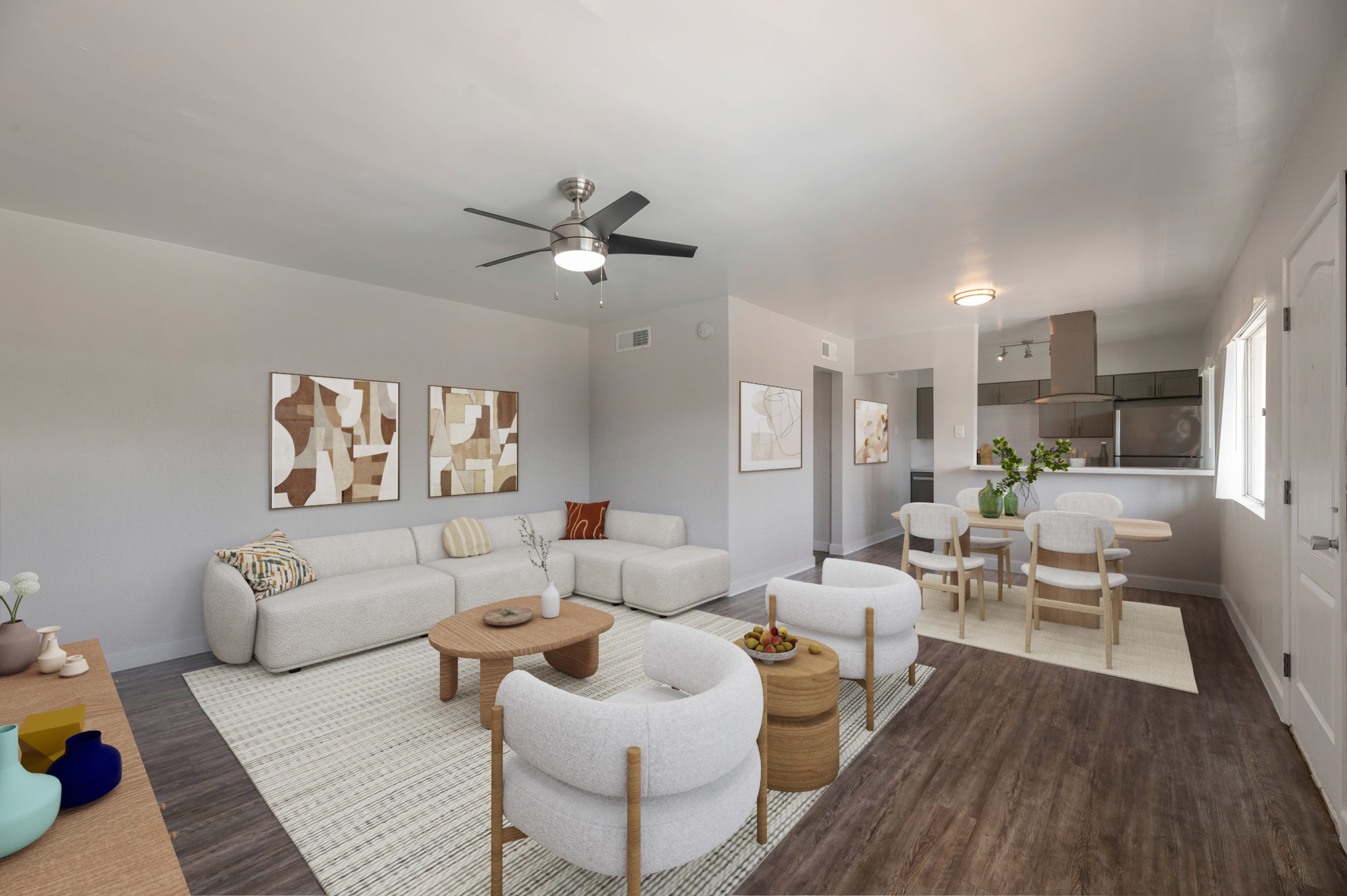 A modern living room featuring a light-colored sectional sofa, two round coffee tables, and a ceiling fan. Abstract artwork adorns the walls. A dining area with a table and chairs is visible, along with a kitchen in the background. Natural light streams in through a window, enhancing the airy atmosphere.
