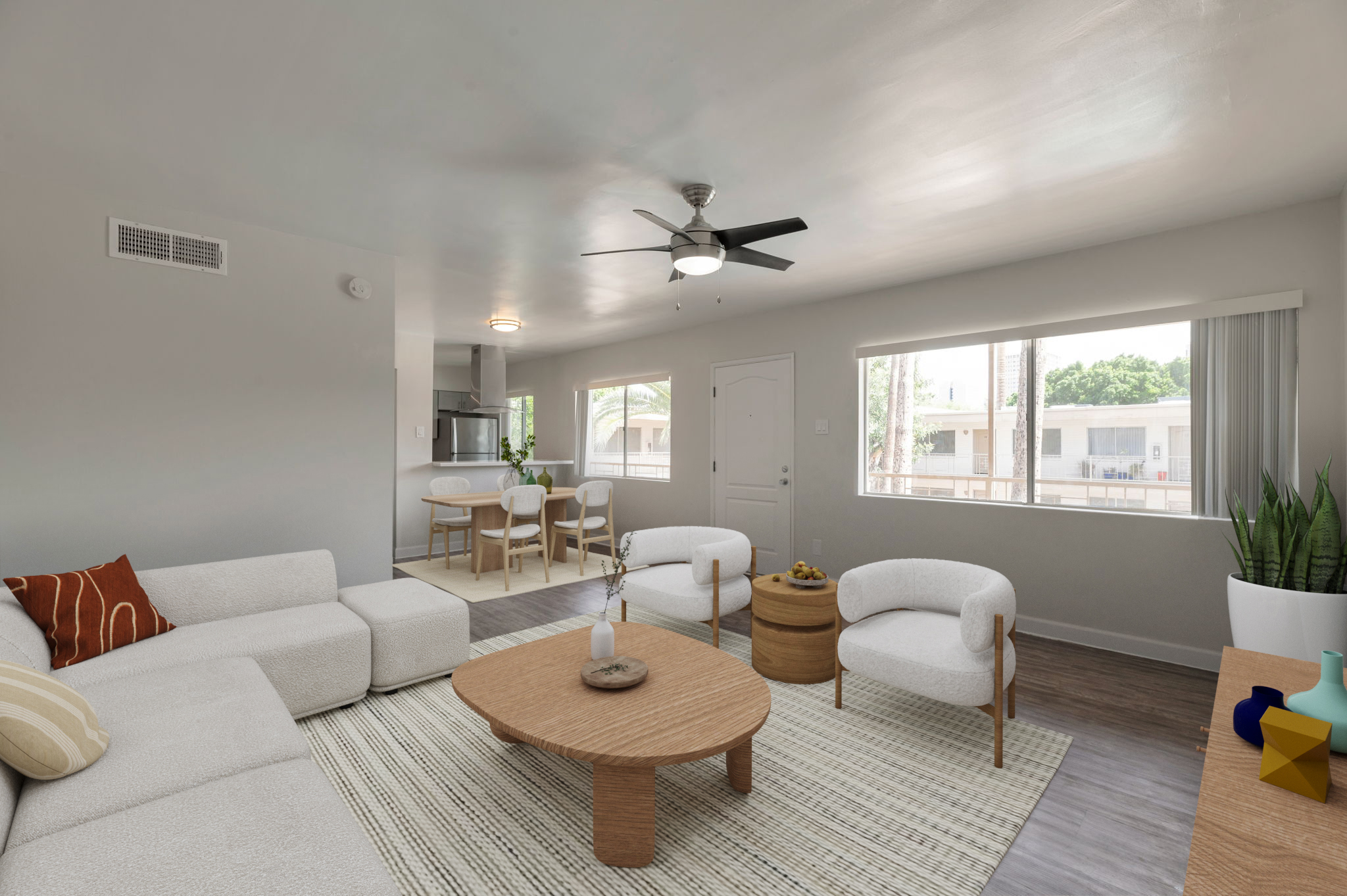 A modern living room featuring a light-colored sofa, two stylish armchairs, and a wooden coffee table. Natural light fills the space through large windows, highlighting the neutral color palette. A small dining area with white chairs and a kitchen can be seen in the background, along with decorative plants and accessories.
