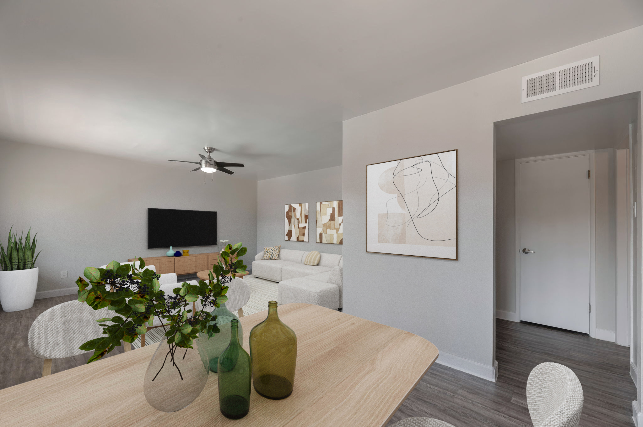 A modern living room featuring a light-colored sofa, a wooden coffee table with decorative vases, and a large wall-mounted TV. Natural light fills the space, complemented by a ceiling fan and minimalist artwork on the walls. A doorway leads to another room, and a green plant adds a touch of nature.