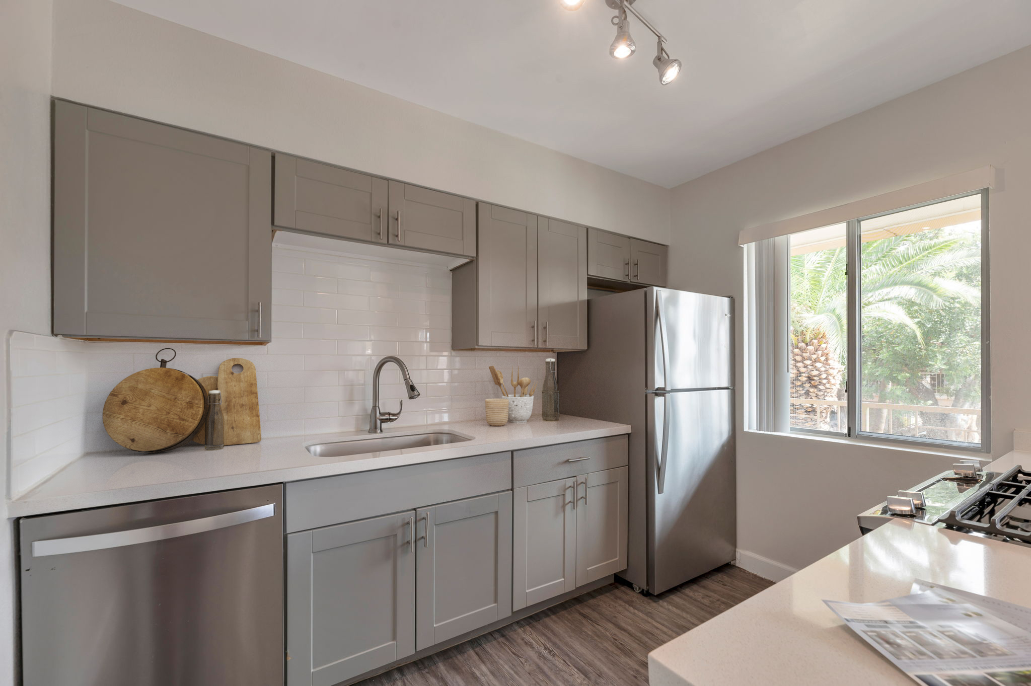 Modern kitchen featuring gray cabinets, a stainless steel refrigerator, and a sleek sink. There's a wooden cutting board on the counter, with a window showcasing greenery outside. The kitchen has a clean, contemporary design with light-colored countertops and natural lighting.