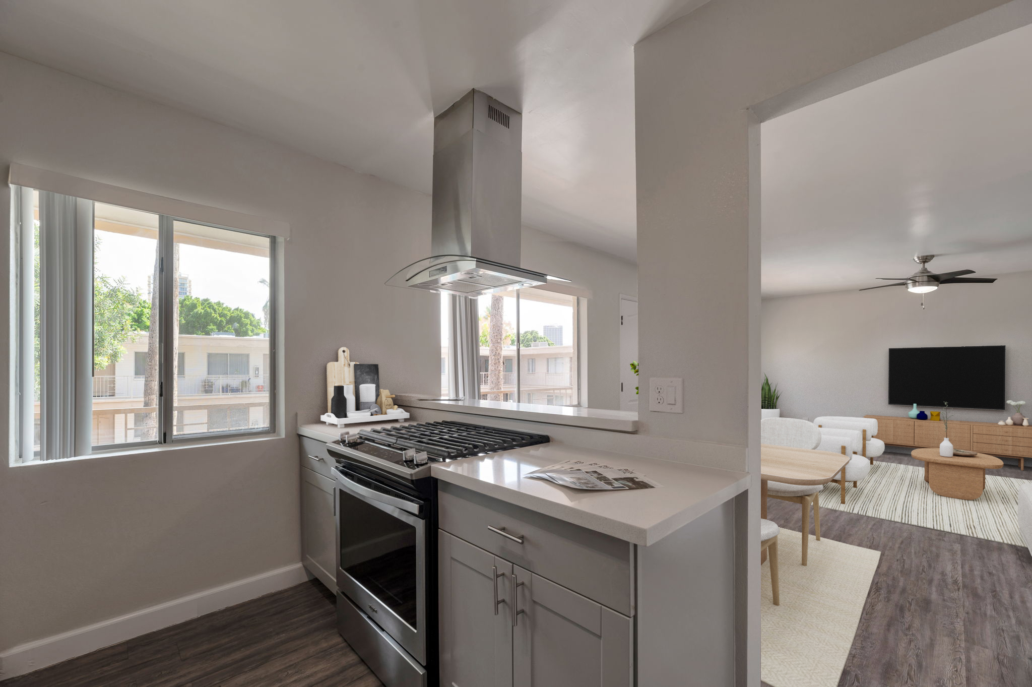 Modern kitchen with a stainless steel gas range and hood, featuring a light-colored countertop and cabinetry. Large windows allow natural light, with a view into an adjacent living area that includes a TV and seating. The overall design is sleek and contemporary.