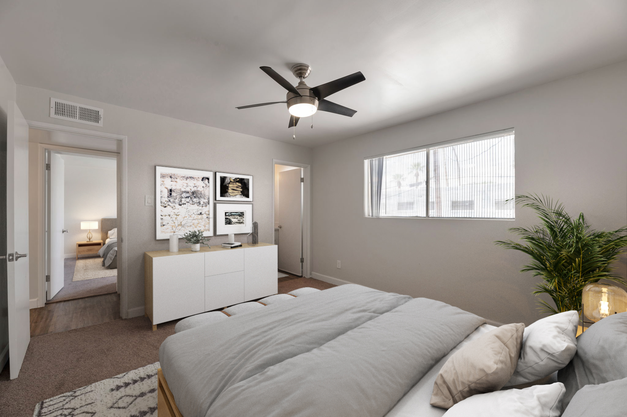A modern bedroom featuring a bed with gray bedding, a white dresser against the wall, and framed art hung above it. A ceiling fan is visible, and there’s a large window allowing natural light. A doorway leads to another room, partially visible, with a cozy atmosphere. A potted plant adds a touch of greenery.