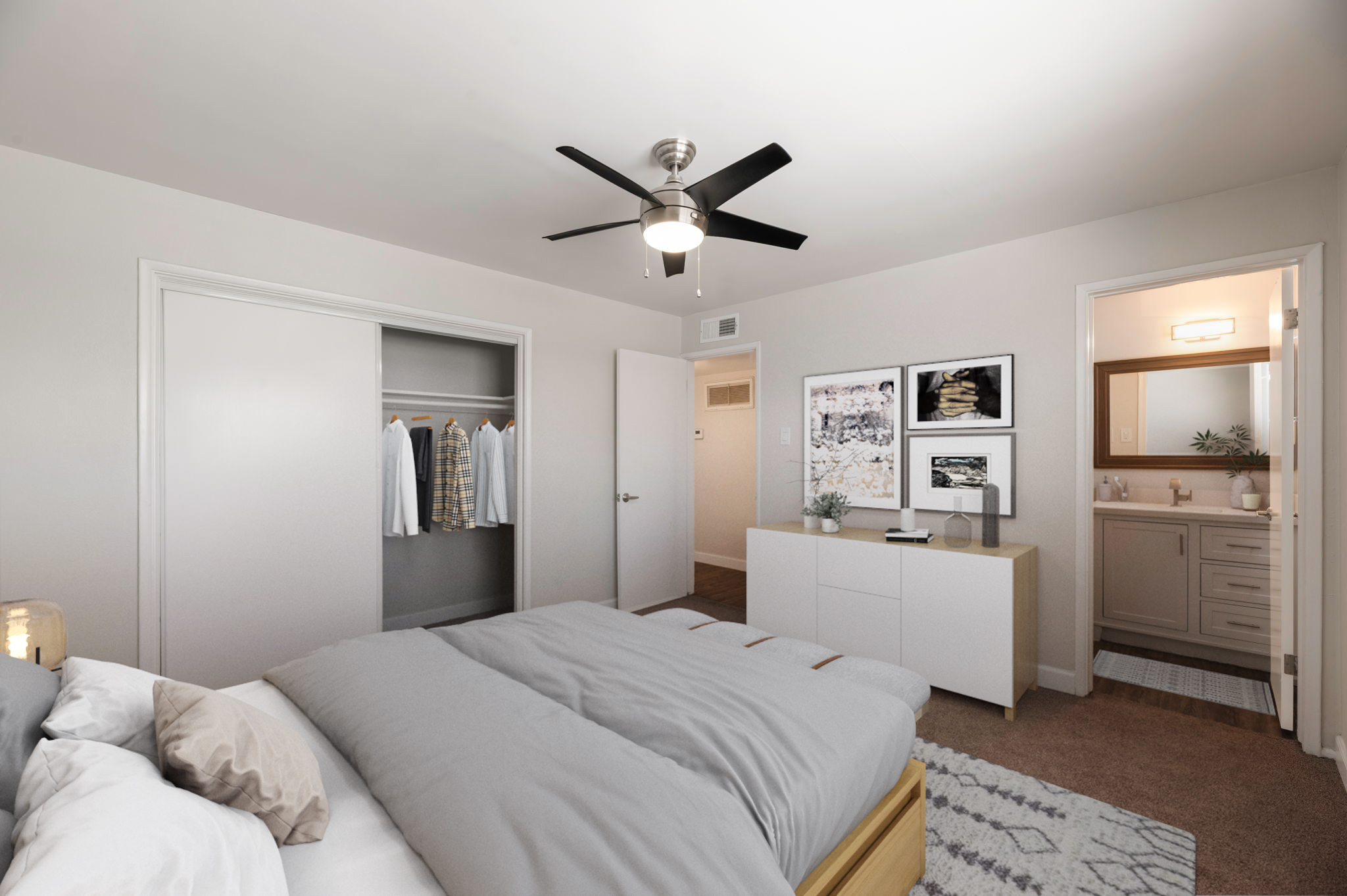 A modern bedroom featuring a neatly made bed with gray bedding, a ceiling fan, and a minimalist dresser. There is an open closet with hanging clothes, and framed artwork on the walls. A bathroom entrance is visible in the background, along with a neutral color palette creating a serene atmosphere.