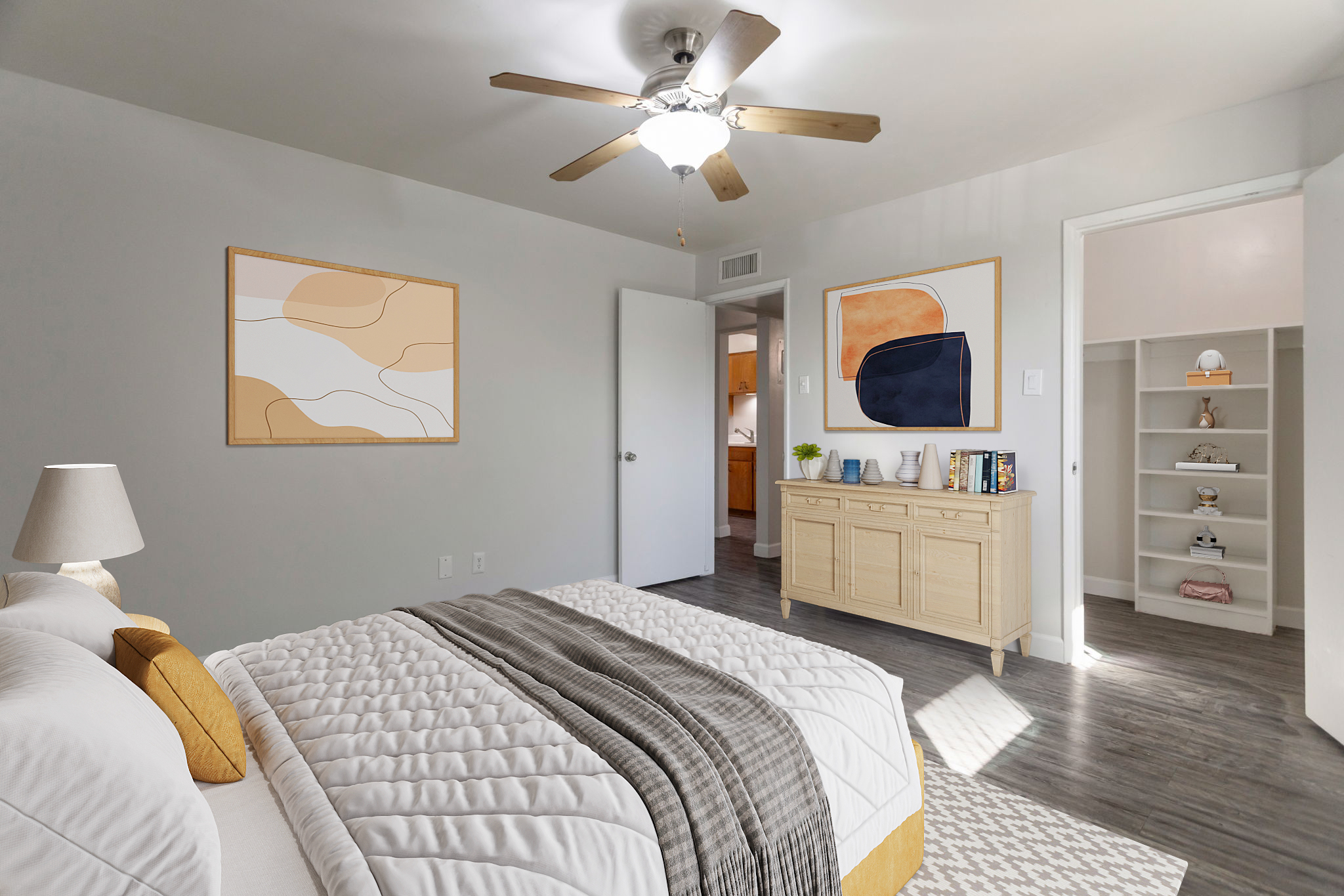 A bright and modern bedroom featuring a cozy bed with white bedding and a yellow accent pillow. A ceiling fan is overhead. Two abstract art pieces adorn the walls. There's a light-colored dresser with books and decor, and a doorway leads to a bathroom area. The room has a clean, minimalist aesthetic with neutral tones.