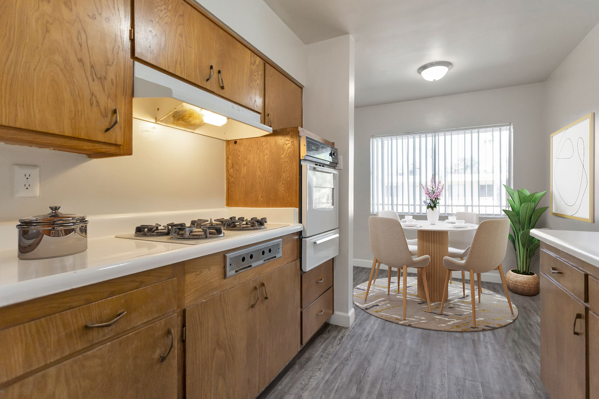 A cozy kitchen featuring wooden cabinets, a gas stove, and a modern oven. A dining area with a round table and two cushioned chairs is visible by a large window, adorned with a vase of flowers. A decorative plant and a circular rug enhance the inviting atmosphere. The space is well-lit and welcoming.