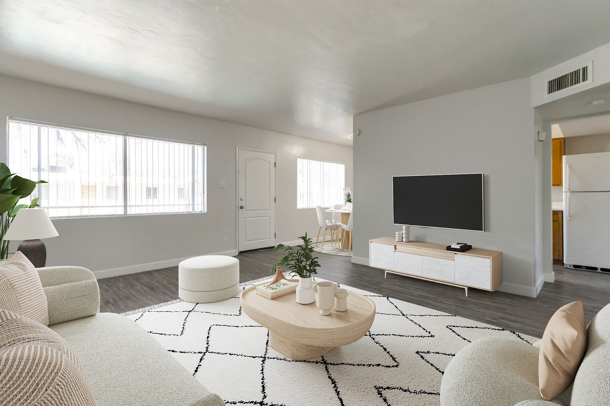 A modern, airy living room featuring a light-colored sofa, a round ottoman, and a coffee table with decorative items. Large windows allow natural light, and a TV is mounted on a light wooden media console. In the background, a dining area with a small table and chairs is visible, enhancing the open layout.