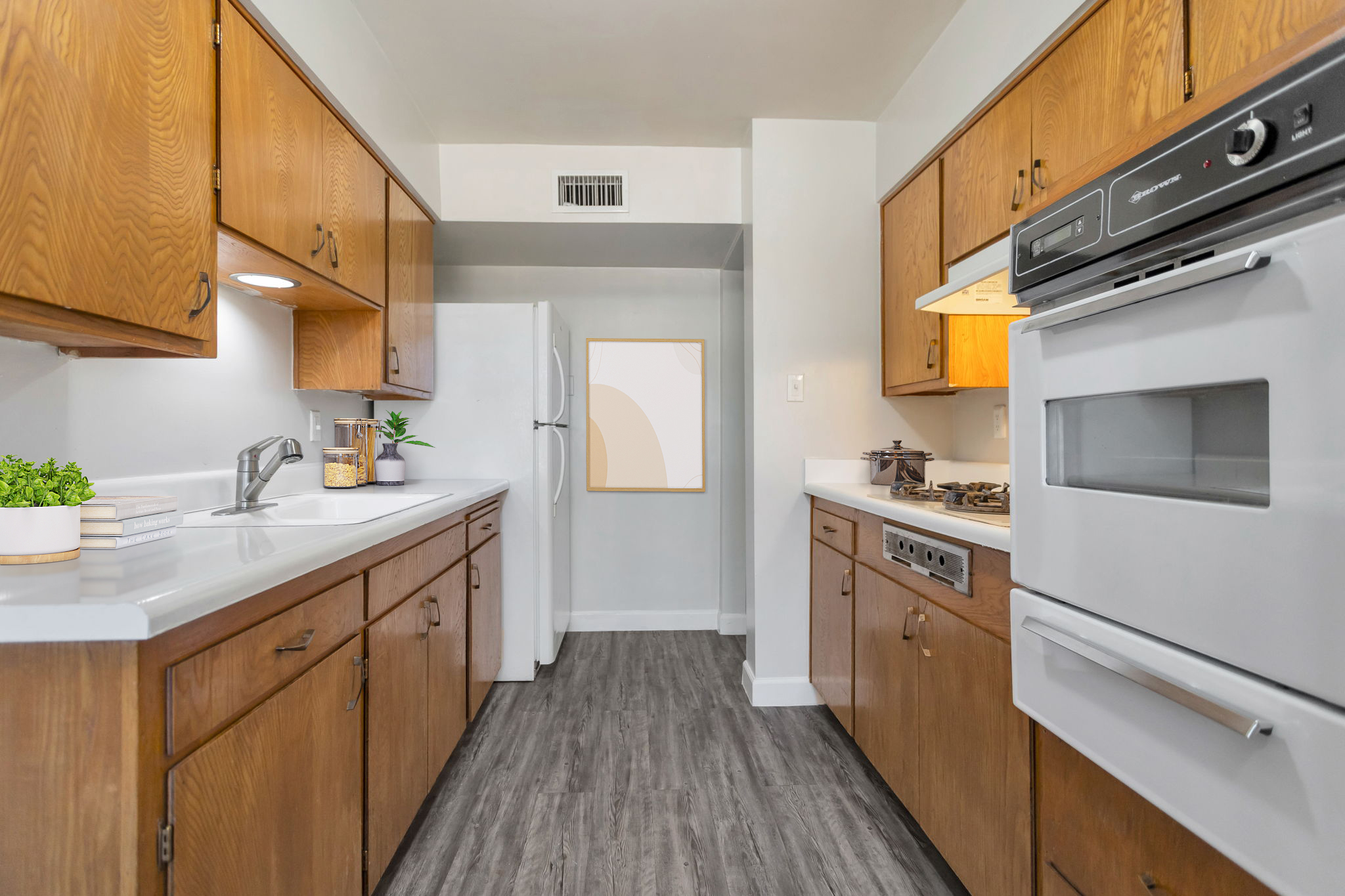 A modern kitchen featuring brown wooden cabinets, white countertops, and stainless steel appliances. The layout includes a refrigerator, a stove, and a sleek sink. Light-colored walls enhance the space, which also has a small decorative plant and a framed artwork on the wall, creating a welcoming atmosphere.