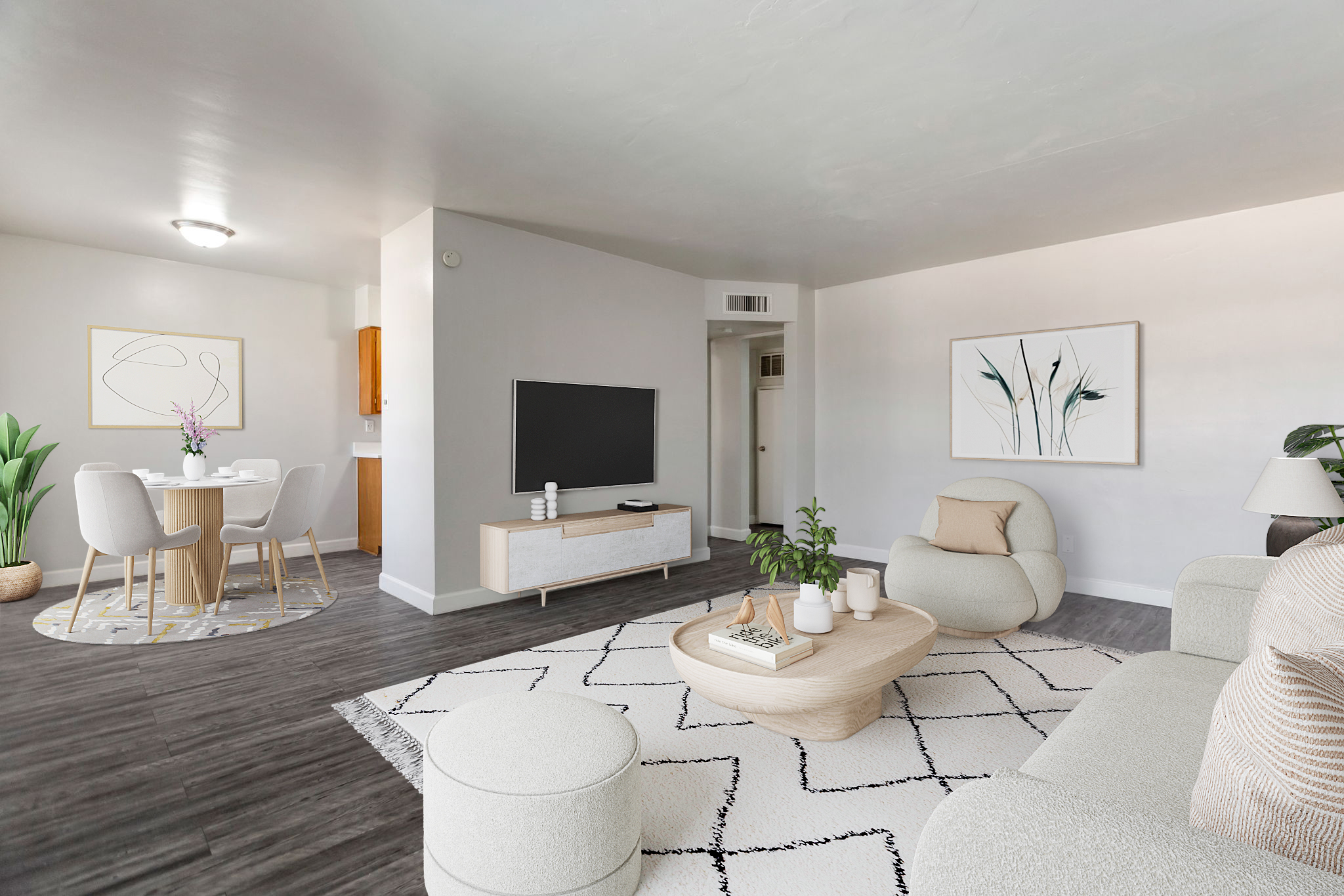 A modern living room featuring a light-colored sofa and armchair, a wooden coffee table with decorative items, and a flat-screen TV on a minimalist stand. There’s a round dining table with chairs in the background, along with a plant and wall art, creating a cozy and inviting atmosphere.