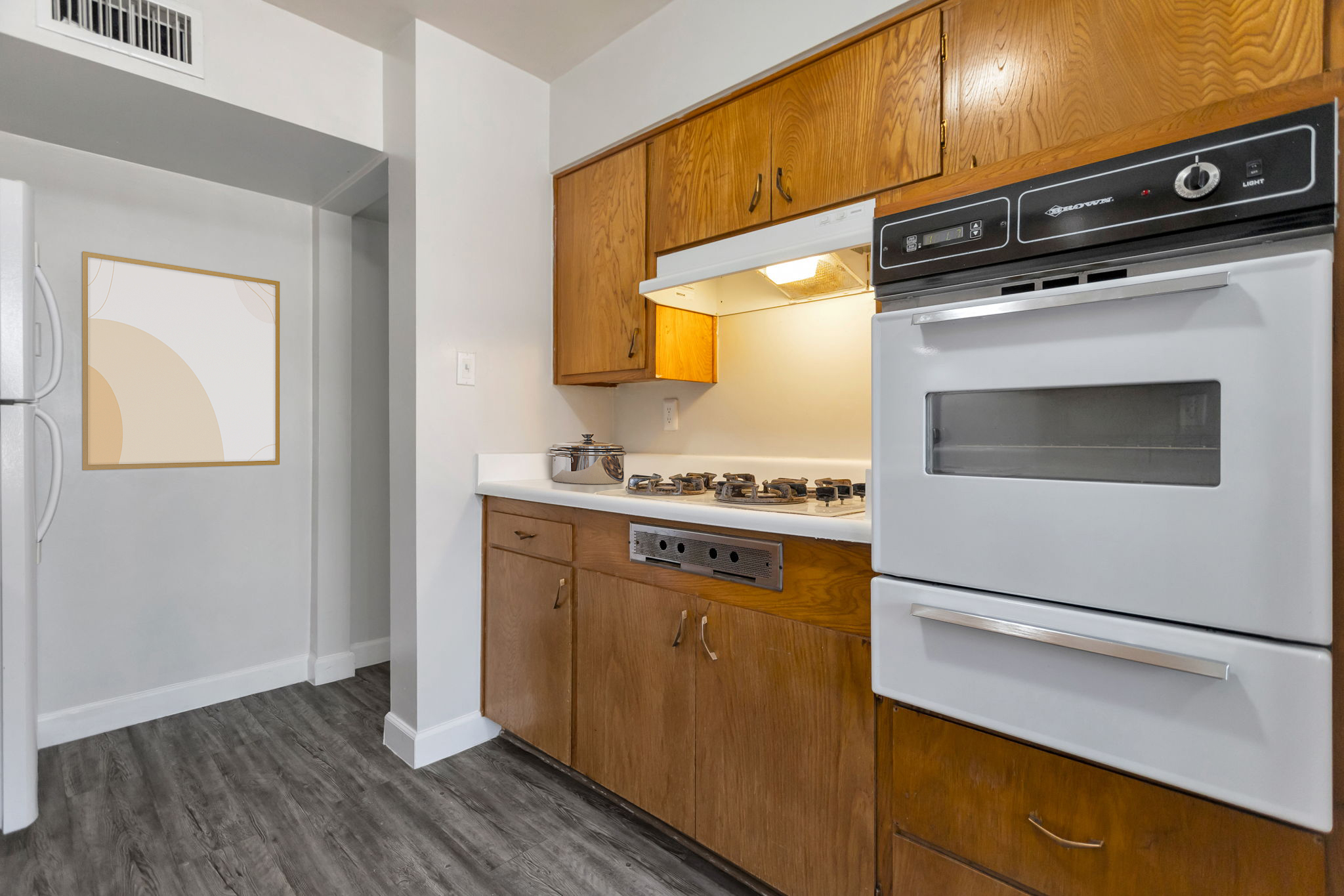 A modern kitchen with wooden cabinets, a white countertop, and stainless steel appliances. The stove has multiple burners, and there is a wall oven above it. The space is well-lit, featuring a blank wall with a minimalist framed artwork. The flooring is a contemporary dark wood style.