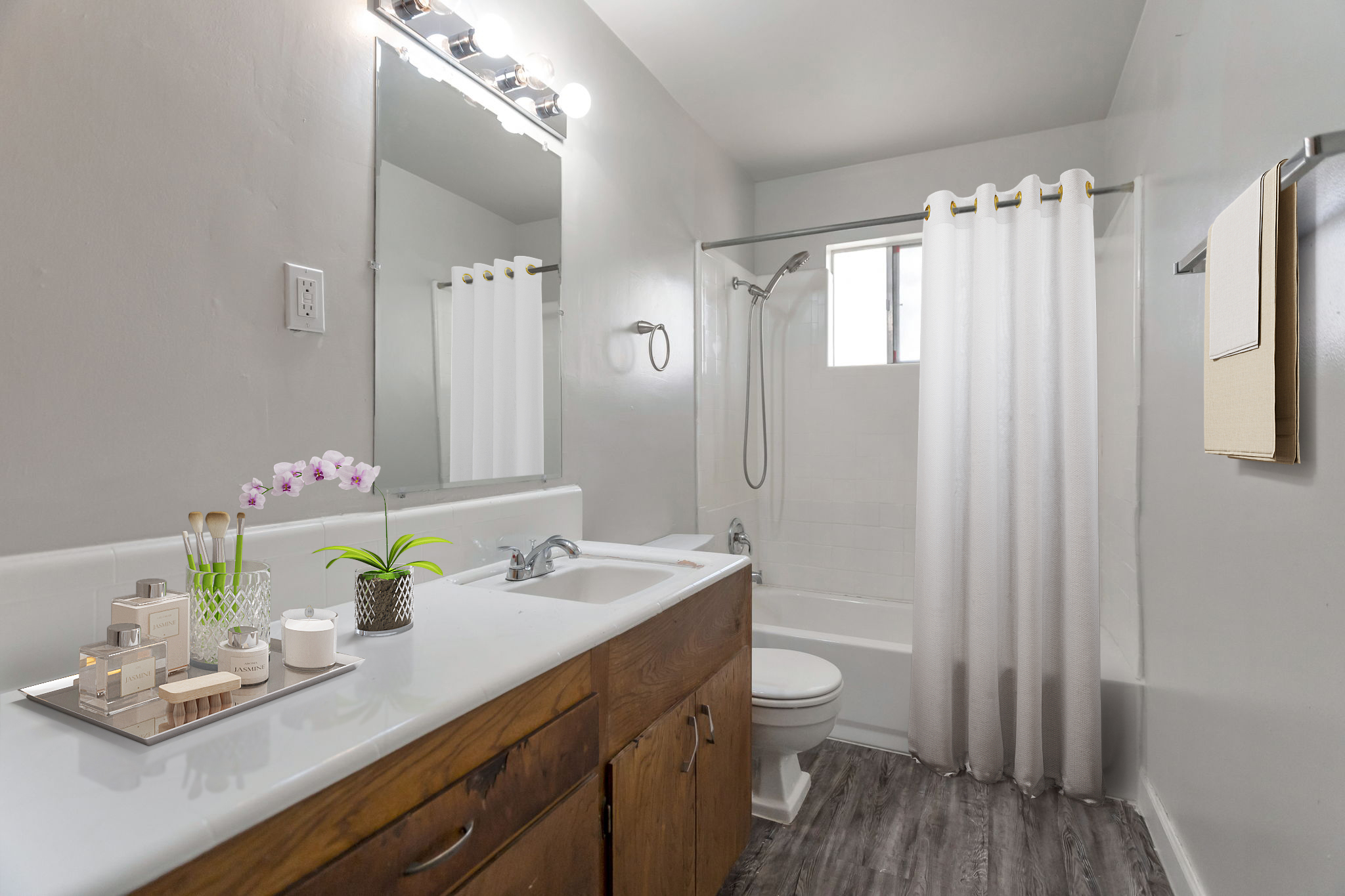 A modern bathroom featuring a white shower curtain, a bathtub with a showerhead, and a sink with wooden cabinetry. The counter displays decorative items like a small plant, candles, and a tray. Light-colored walls and a window create a bright, airy atmosphere.