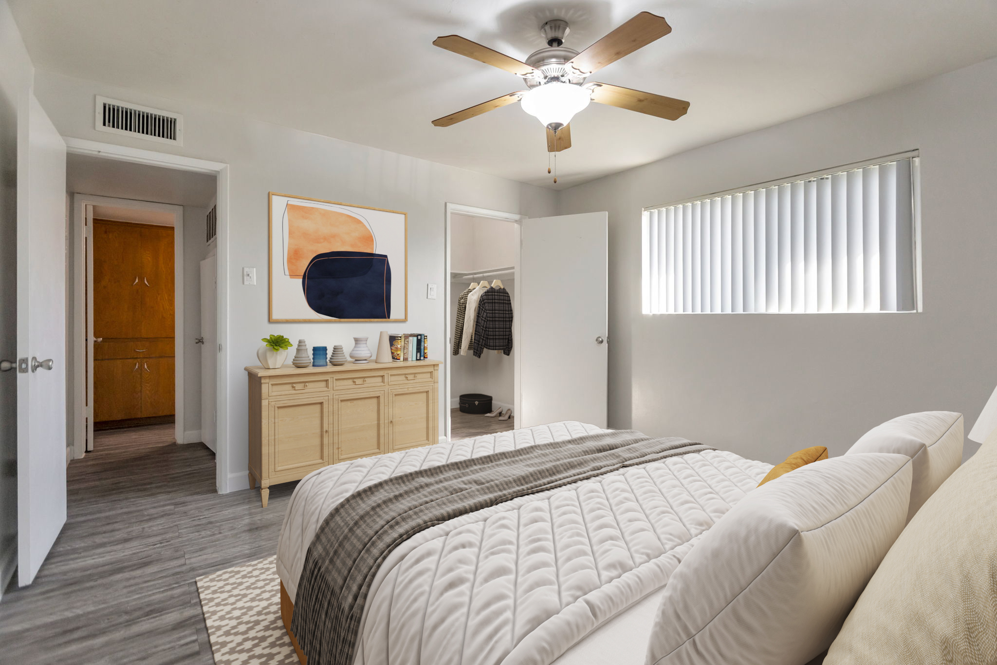 A well-lit bedroom featuring a white bed with textured linens, a wooden dresser, and a ceiling fan. A closet door is slightly open, showing hanging clothes. There is a window with vertical blinds, and a hallway leading to another room is visible. A piece of abstract art adorns the wall above the dresser.