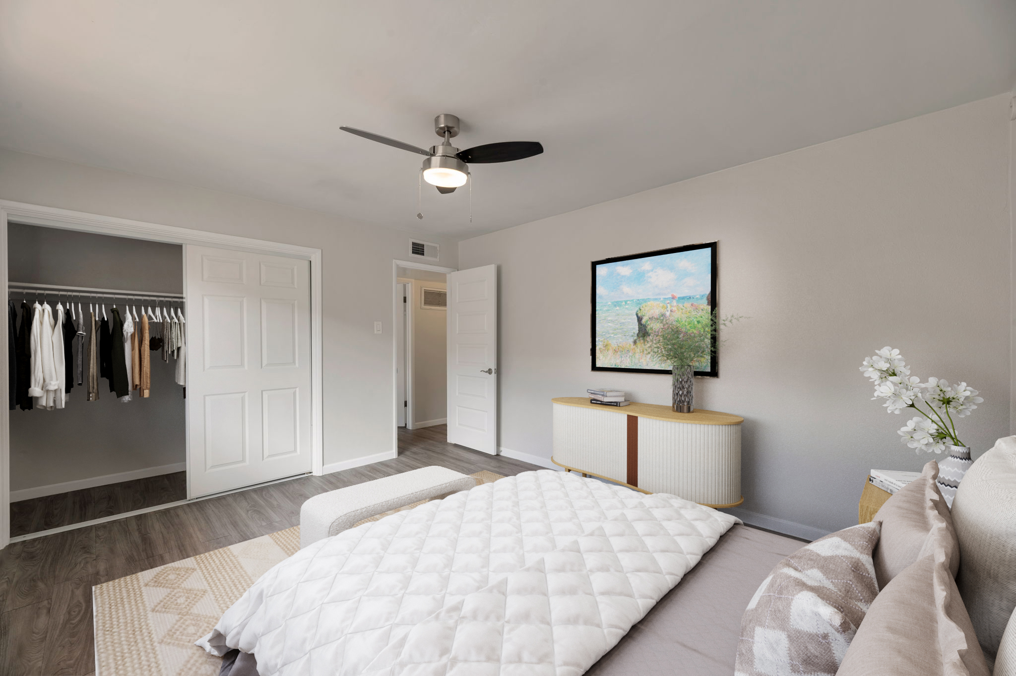 A modern bedroom featuring a neatly made bed with a quilted cover, a small ottoman, and a stylish dresser. A large closet with clothes is visible, and a framed painting hangs on the wall above a vase with flowers. The room has light-colored walls and a ceiling fan, creating a bright and airy atmosphere.