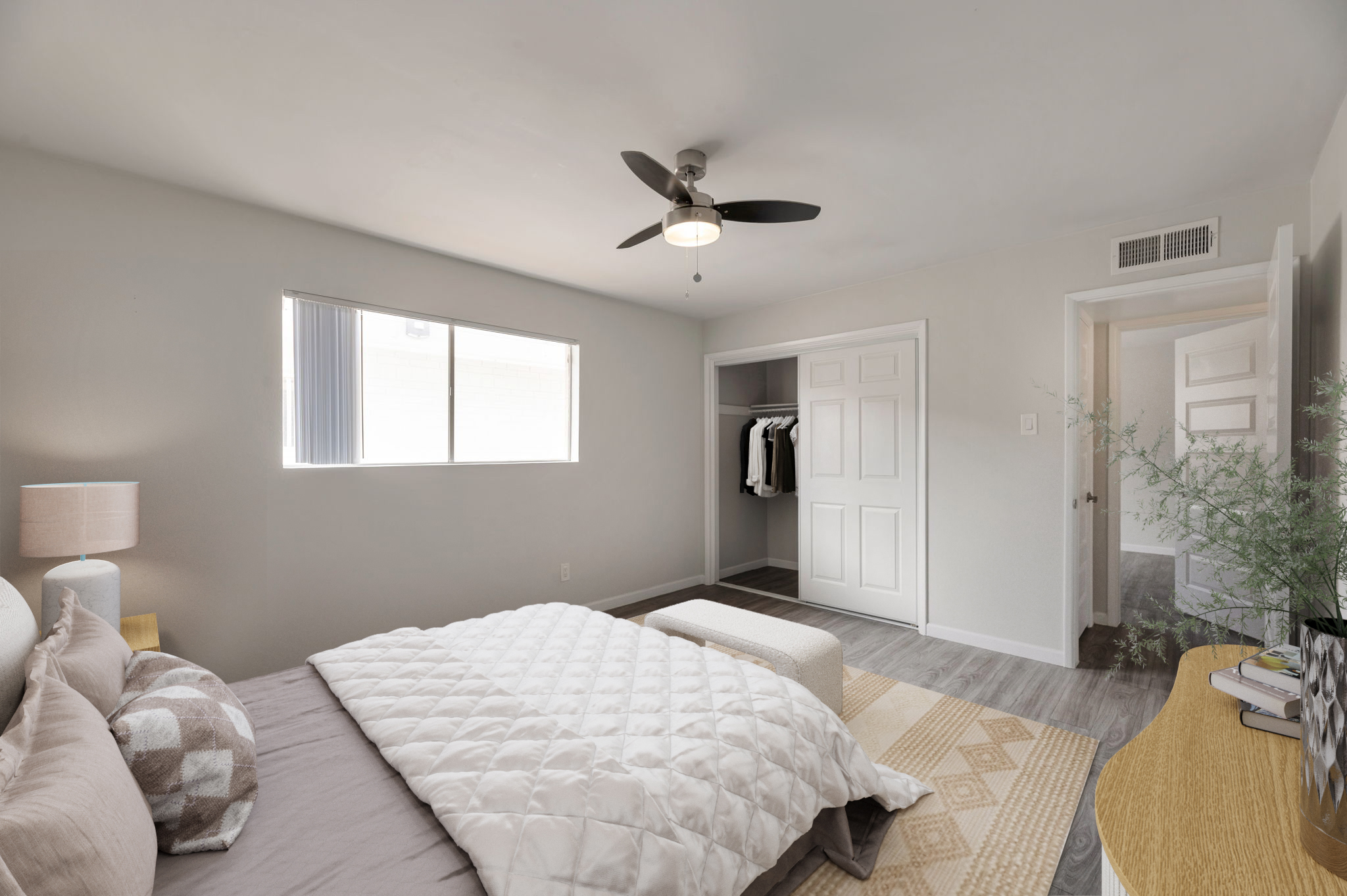 A cozy bedroom featuring a large window letting in natural light, a neatly made bed with a white quilt, a modern ceiling fan, and a wooden side table with a lamp. A closet can be seen with clothes hanging, and a doorway leads to another room. The decor is minimalist and inviting.