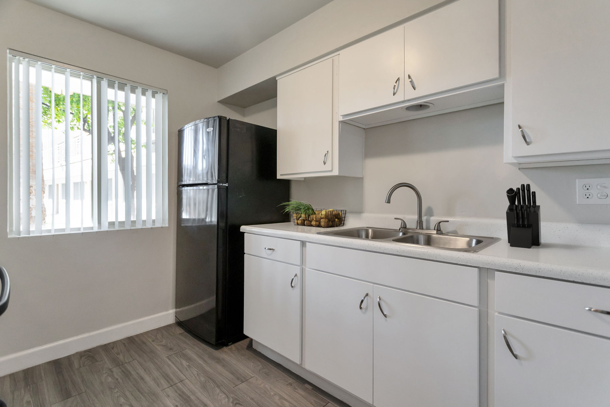 Modern kitchen featuring white cabinetry, a double sink, and a black refrigerator. Natural light filters through white vertical blinds. A small potted plant is placed next to the sink, and kitchen utensils are arranged neatly in a container on the counter. The floor has a wood-like appearance.