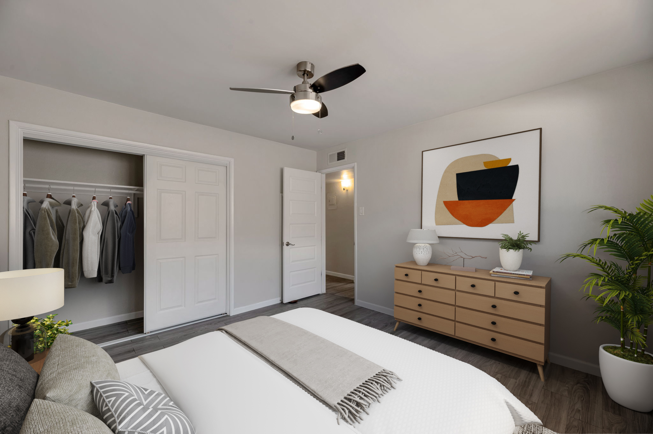 A well-decorated bedroom featuring a cozy bed with gray and white bedding, an abstract art piece on the wall, a wooden dresser, a ceiling fan, and a potted plant. A closet with neatly hung clothes is visible, and a doorway leads to another part of the home. The room has a neutral color palette with soft lighting.
