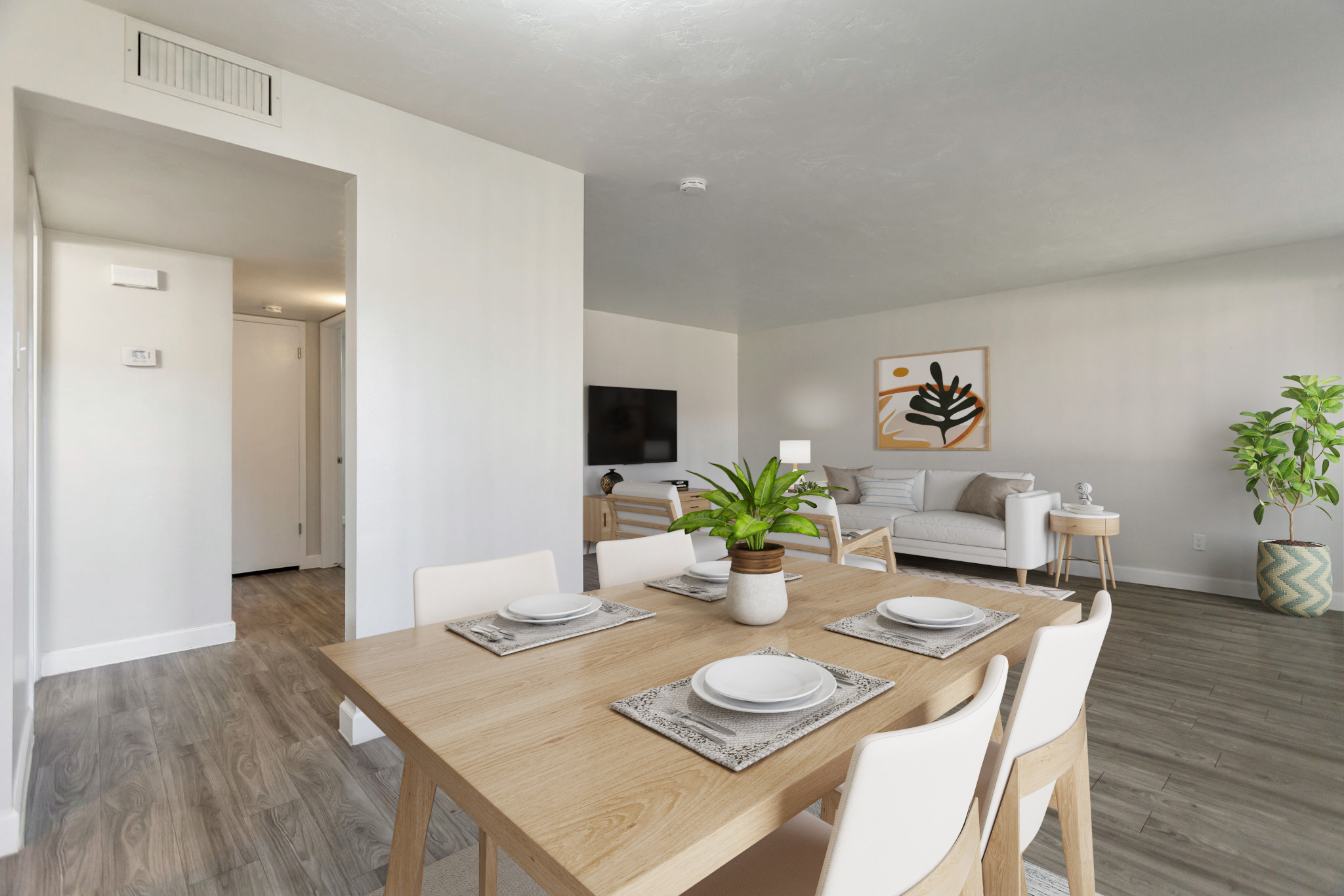 A modern, bright dining area featuring a wooden table set with white plates and napkins, surrounded by white chairs. In the background, a cozy living space is visible with a light-colored sofa, a TV mounted on the wall, and a decorative plant. The walls are painted in neutral tones.