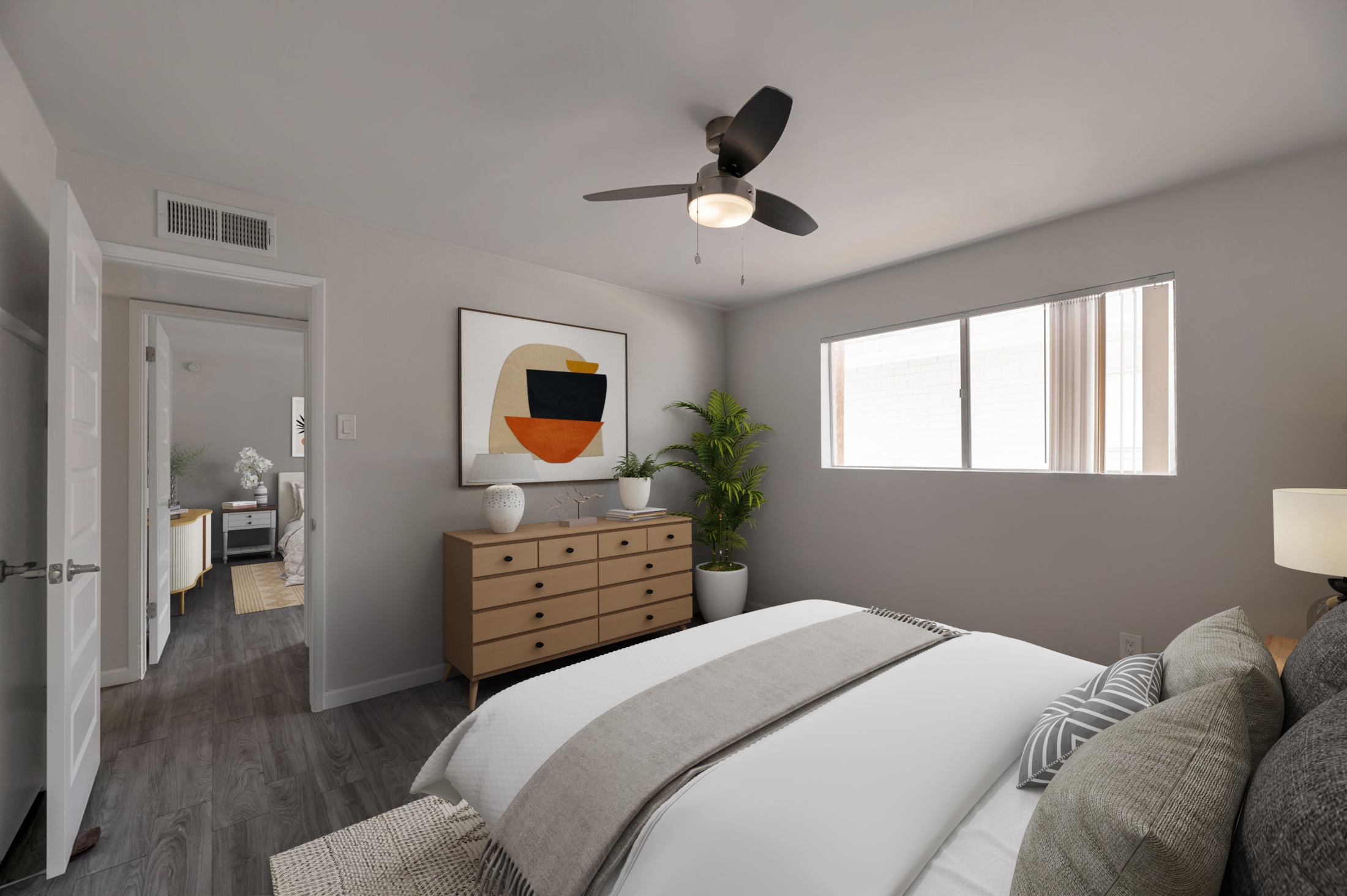 A modern bedroom featuring a double bed with gray and white bedding. There's a wooden dresser, a ceiling fan, and a large window allowing natural light. A framed abstract art piece hangs on the wall, and a potted plant adds a touch of greenery. The room has a minimalist and cozy aesthetic.