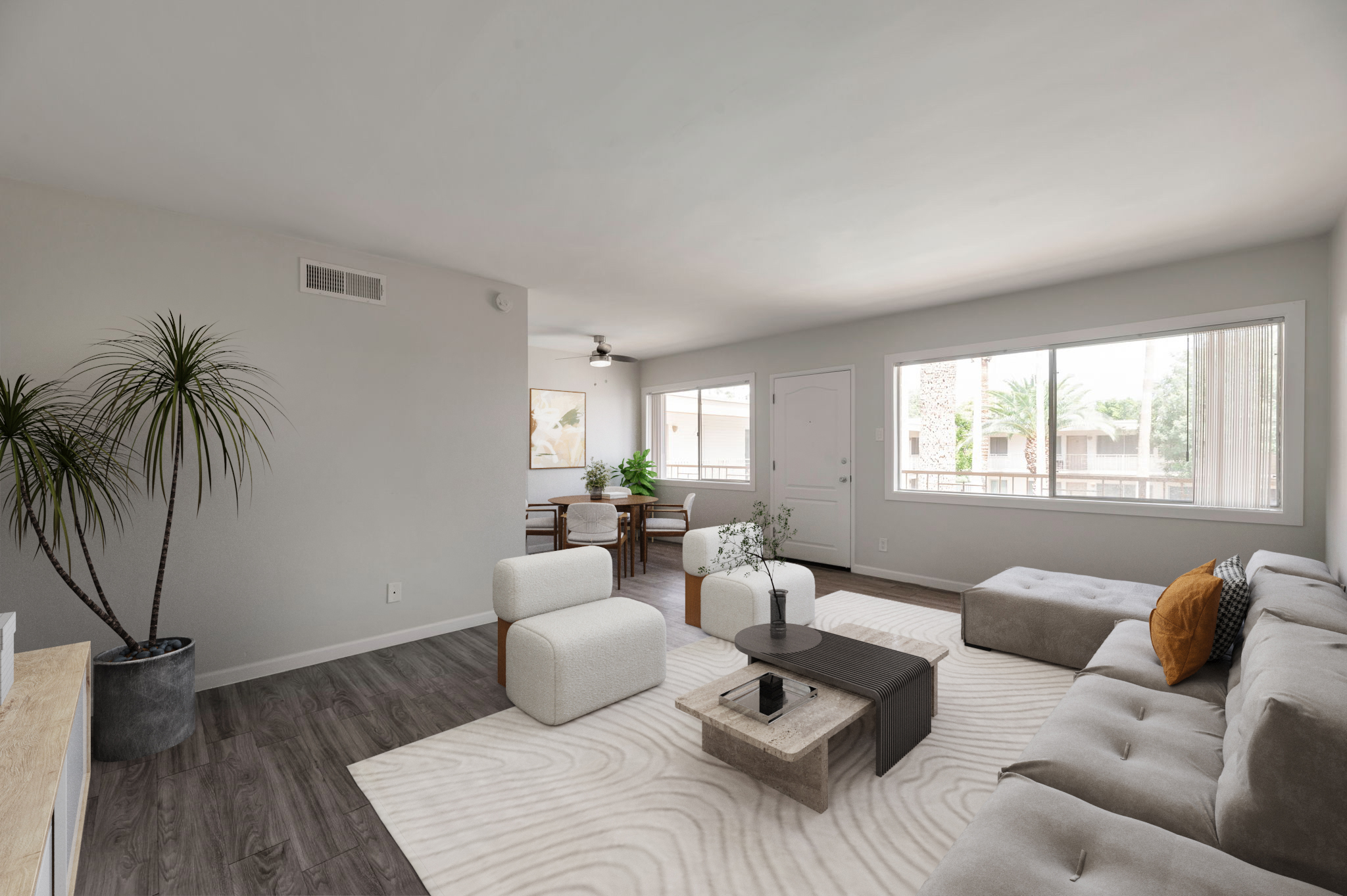 A modern, airy living room featuring a light-colored sofa with decorative cushions, a coffee table with plants, and a large area rug. Large windows allow natural light to fill the space, which includes a dining area with wooden chairs and a table in the background.